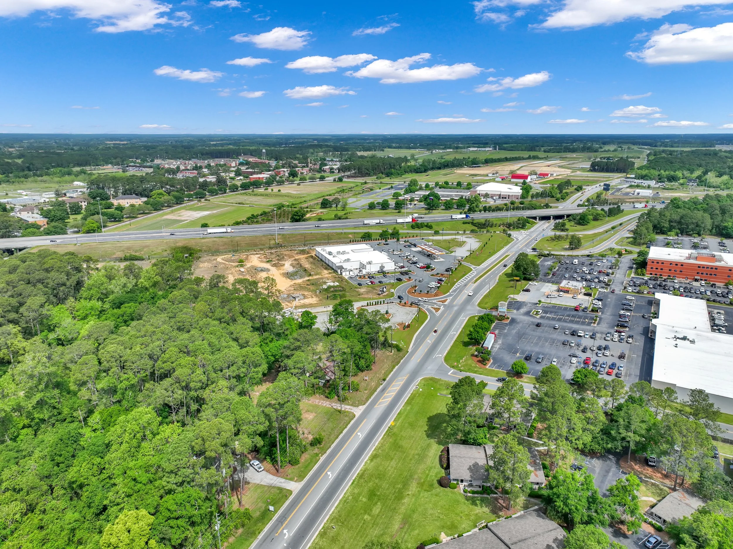 Aerial view of a suburban area showing roads, parking lots, commercial buildings, and green wooded areas under a partly cloudy sky.
