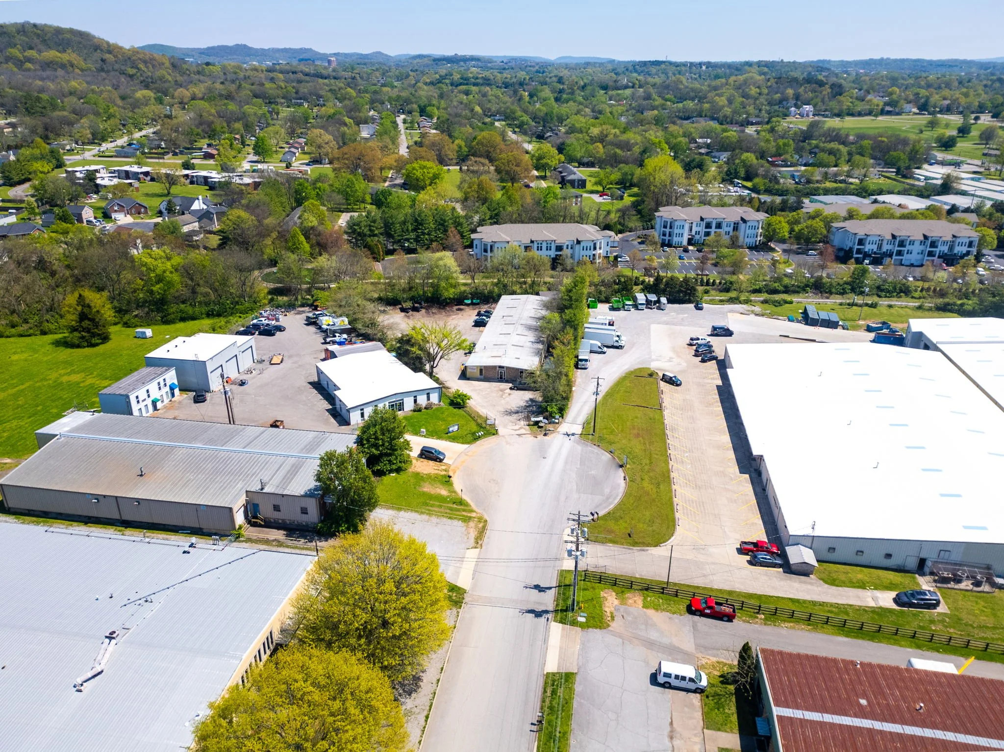 Aerial view of an industrial area with warehouses, commercial buildings, and parking lots surrounded by trees and residential neighborhoods in the background.