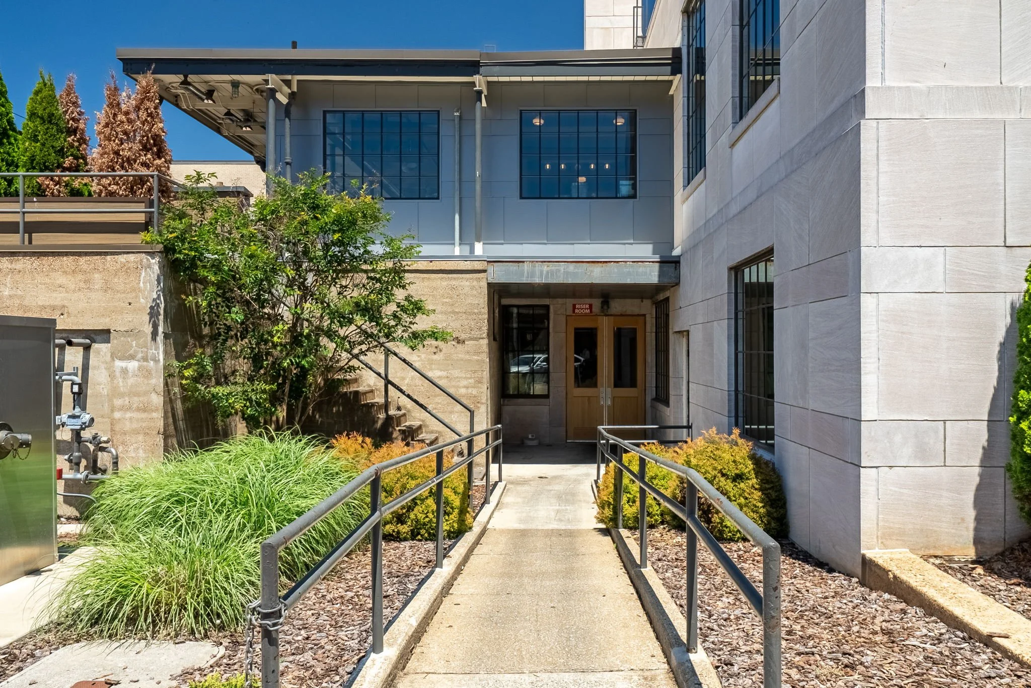 A modern building with large glass windows, a pathway with handrails, and surrounding greenery including bushes and trees, on a sunny day with a bright blue sky.