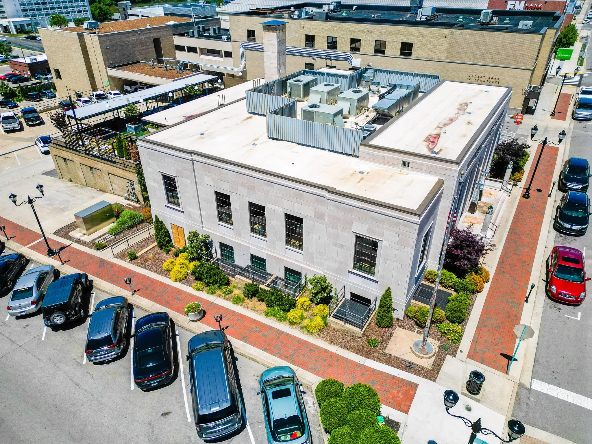 An aerial view of a city building rooftop with HVAC units, parking lots filled with cars, surrounding sidewalks, street lamps, and nearby buildings.