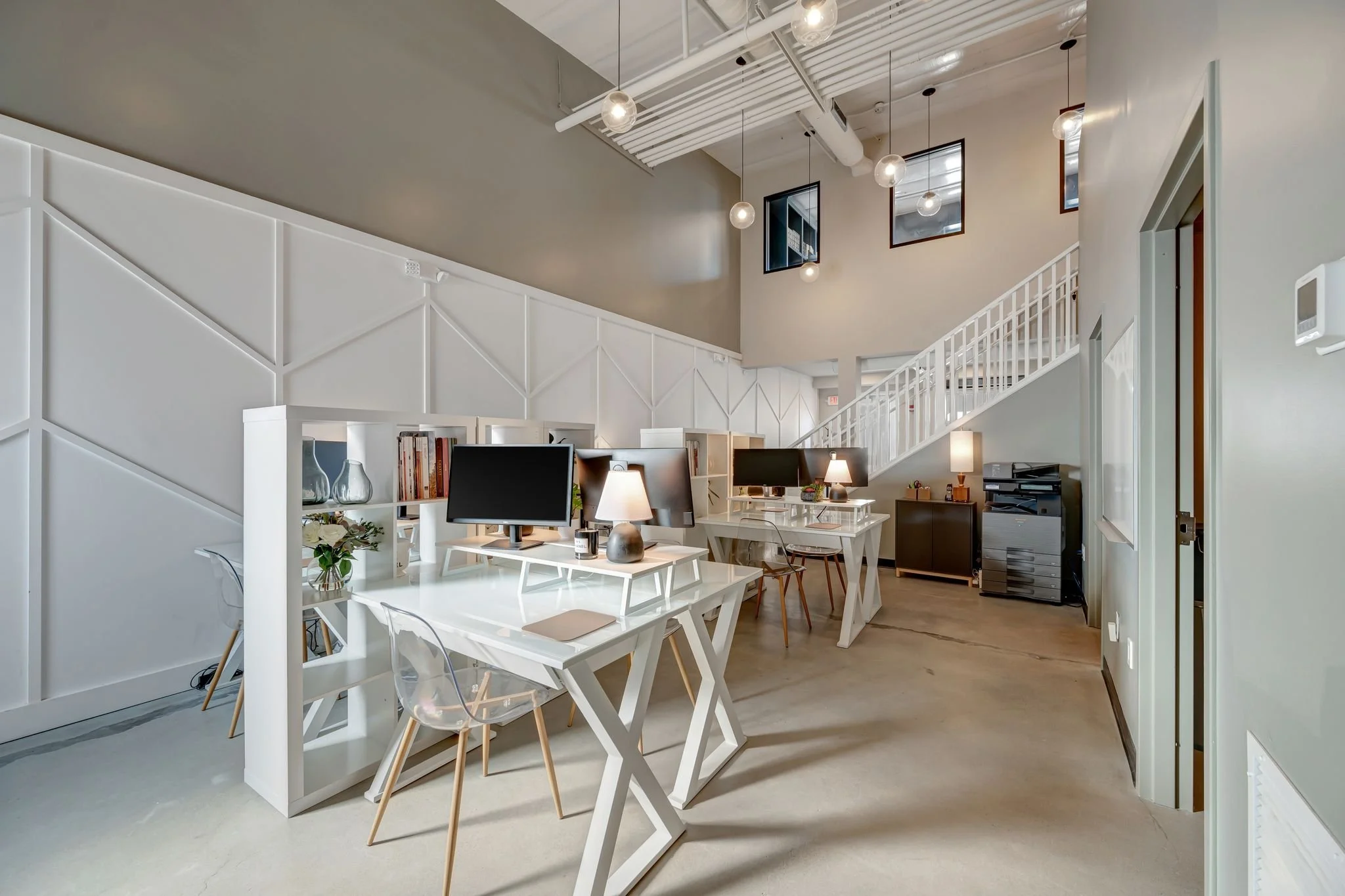 Modern office space with white desks, computer monitors, lamps, and transparent chairs, along with a staircase and decorative lighting fixtures.