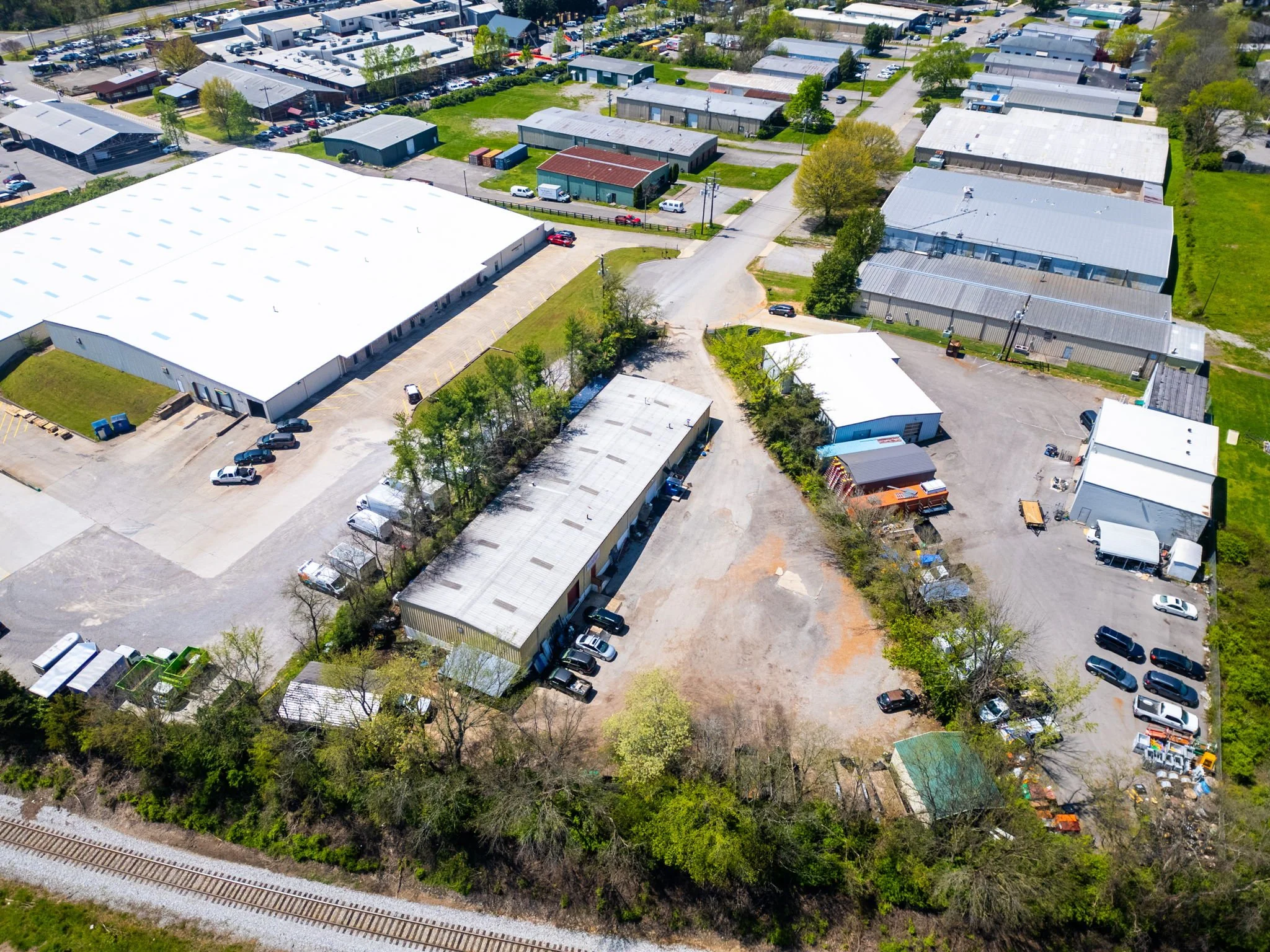 Aerial view of an industrial area with large warehouse buildings, smaller structures, parking lots, and surrounding greenery, near a railroad track.
