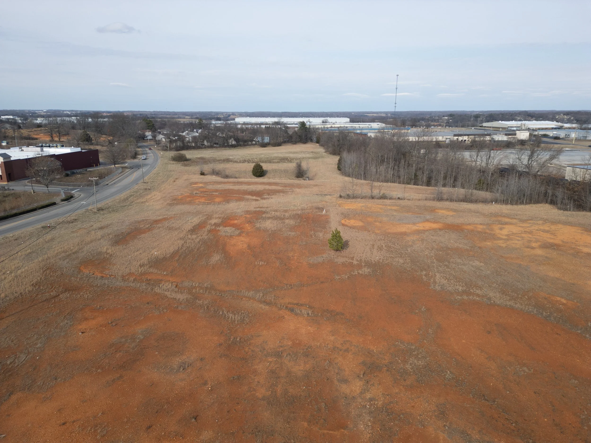 An aerial view of undeveloped land with patches of reddish-brown soil, grassy areas, and a few scattered trees. Near a road on the left, there are some industrial buildings and warehouse-like structures in the background.