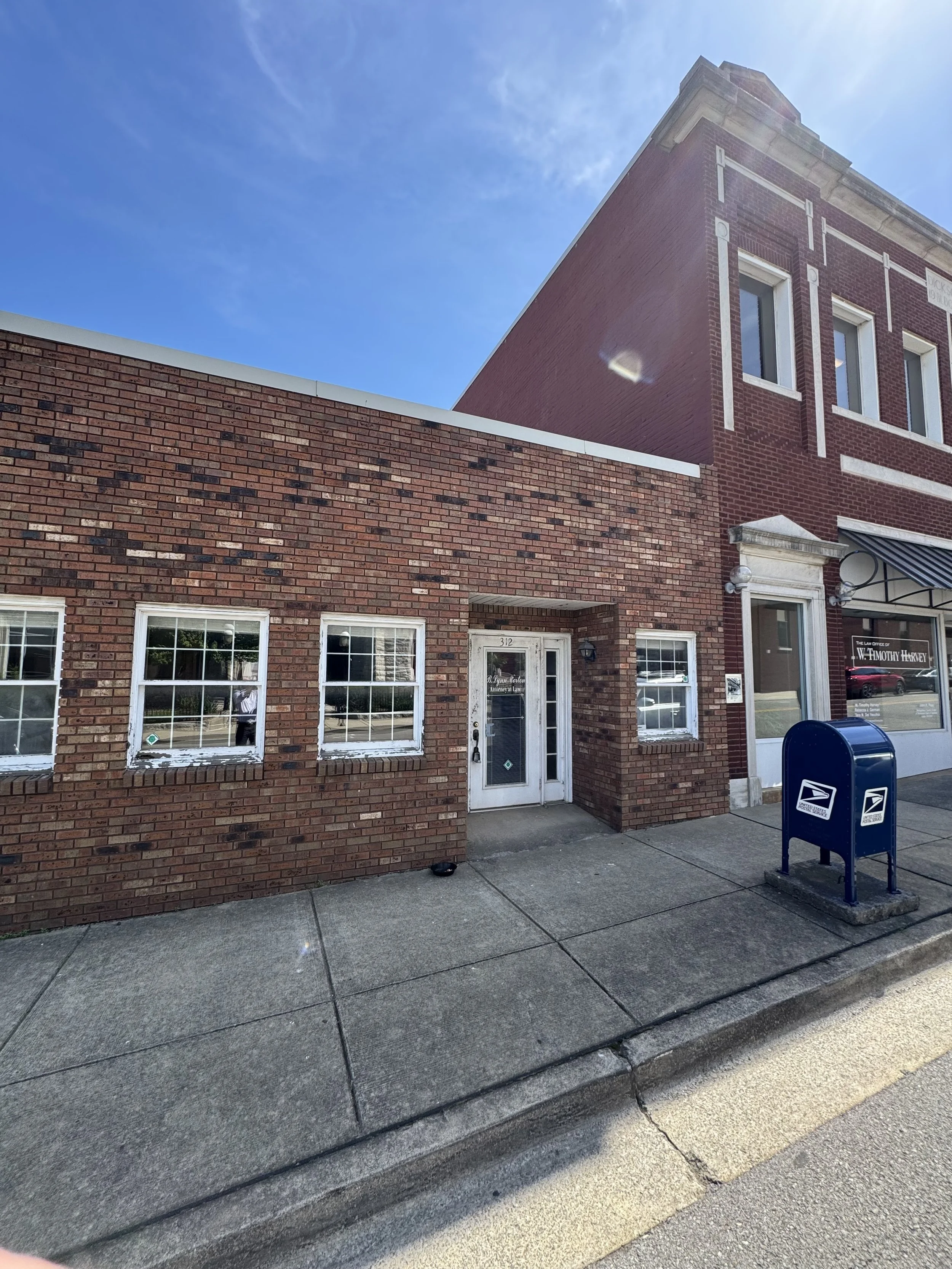 Street view of a small brick building with four windows and a door, next to a larger brick building with a storefront and mailbox, under a sunny blue sky.