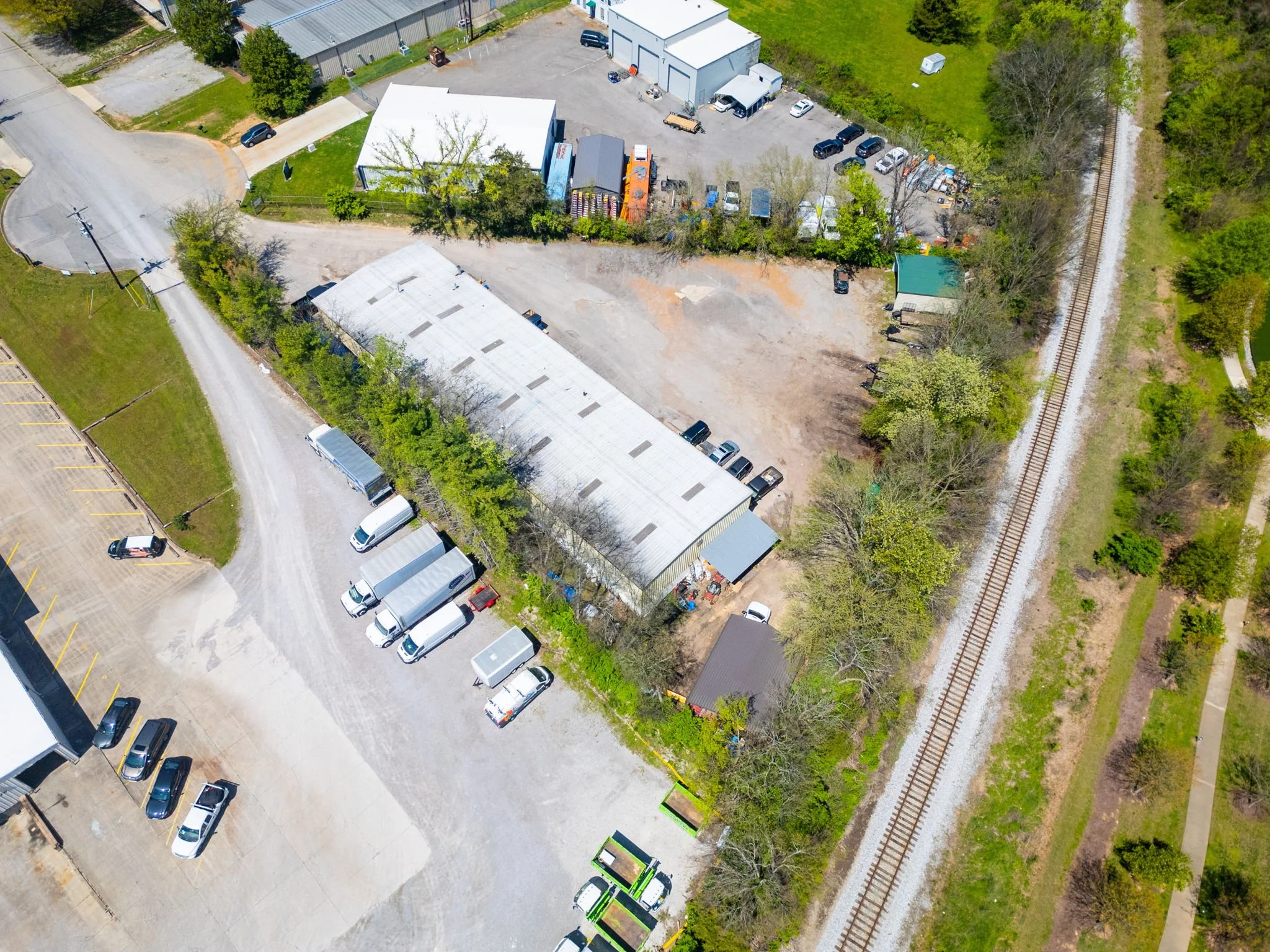 An aerial view of a small industrial or commercial property with parking lots, storage buildings, trees, and a nearby railway track.