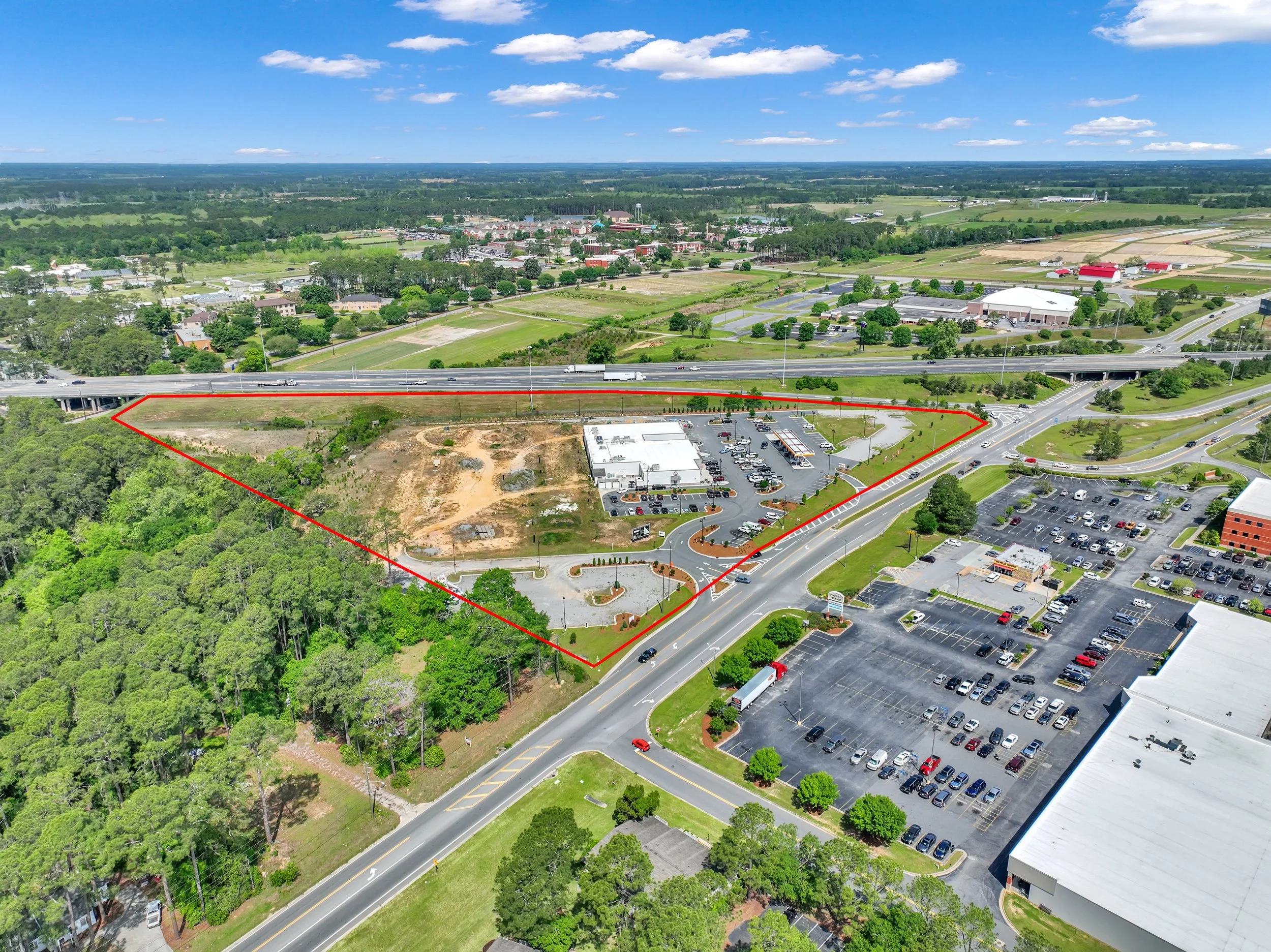 Aerial view of a commercial area with parking lots, a shopping center, nearby wooded area, and a highway running across the top.