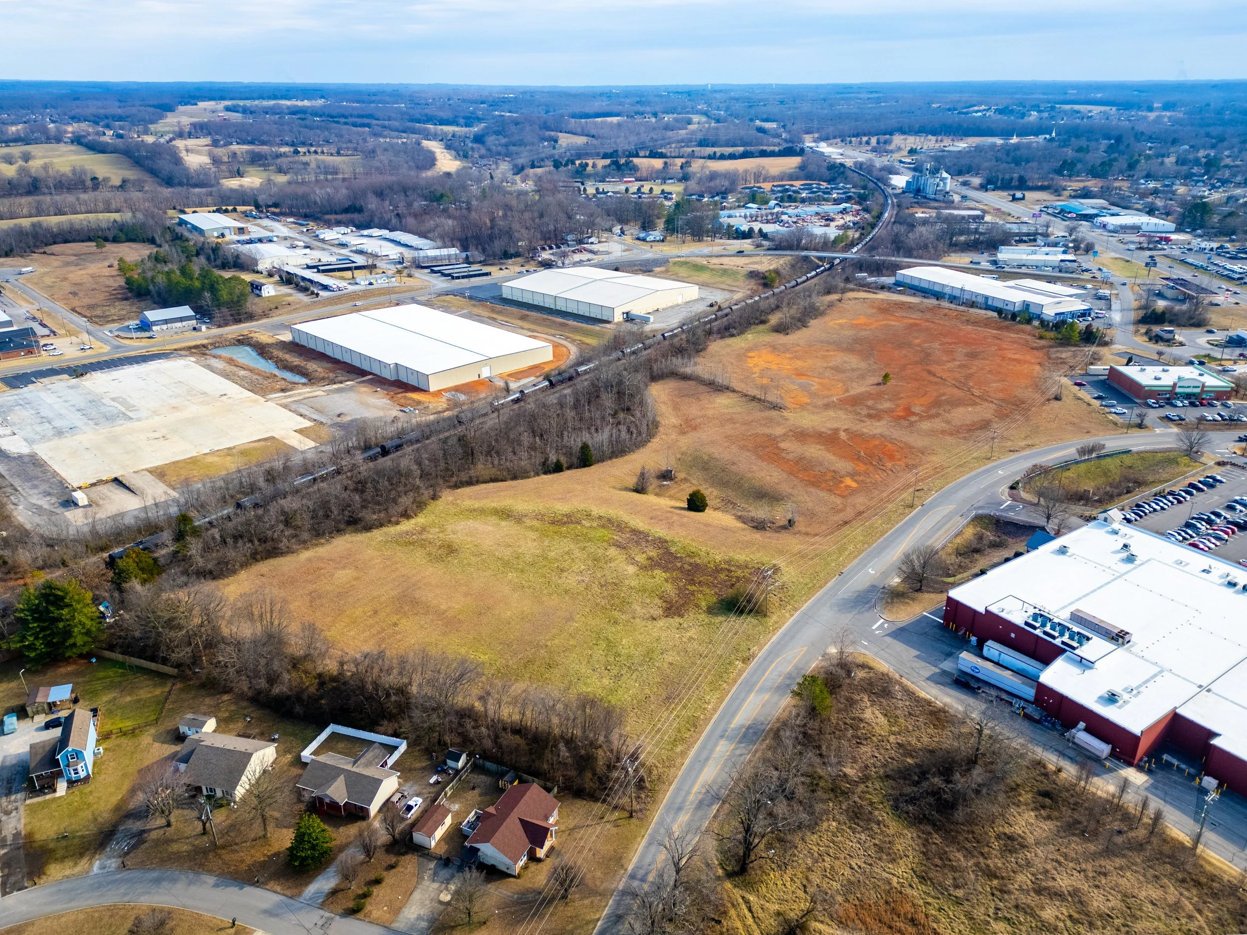 Aerial view of a commercial area with empty lots, warehouses, and residential houses, with roads and parking lots visible.
