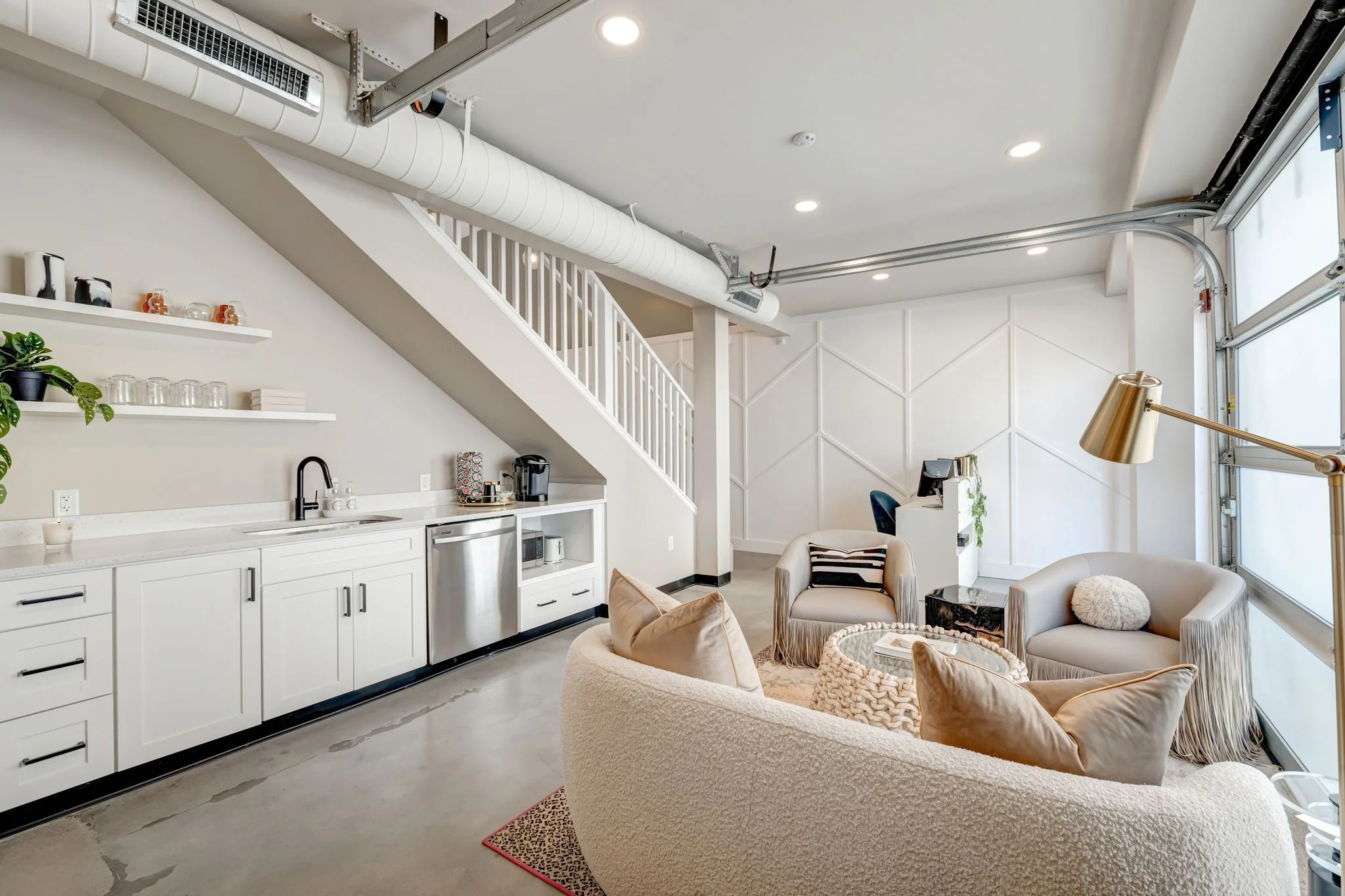 Cozy modern lounge with white cabinetry, seating area with beige and charcoal chairs, gold floor lamp, and windowed garage door.
