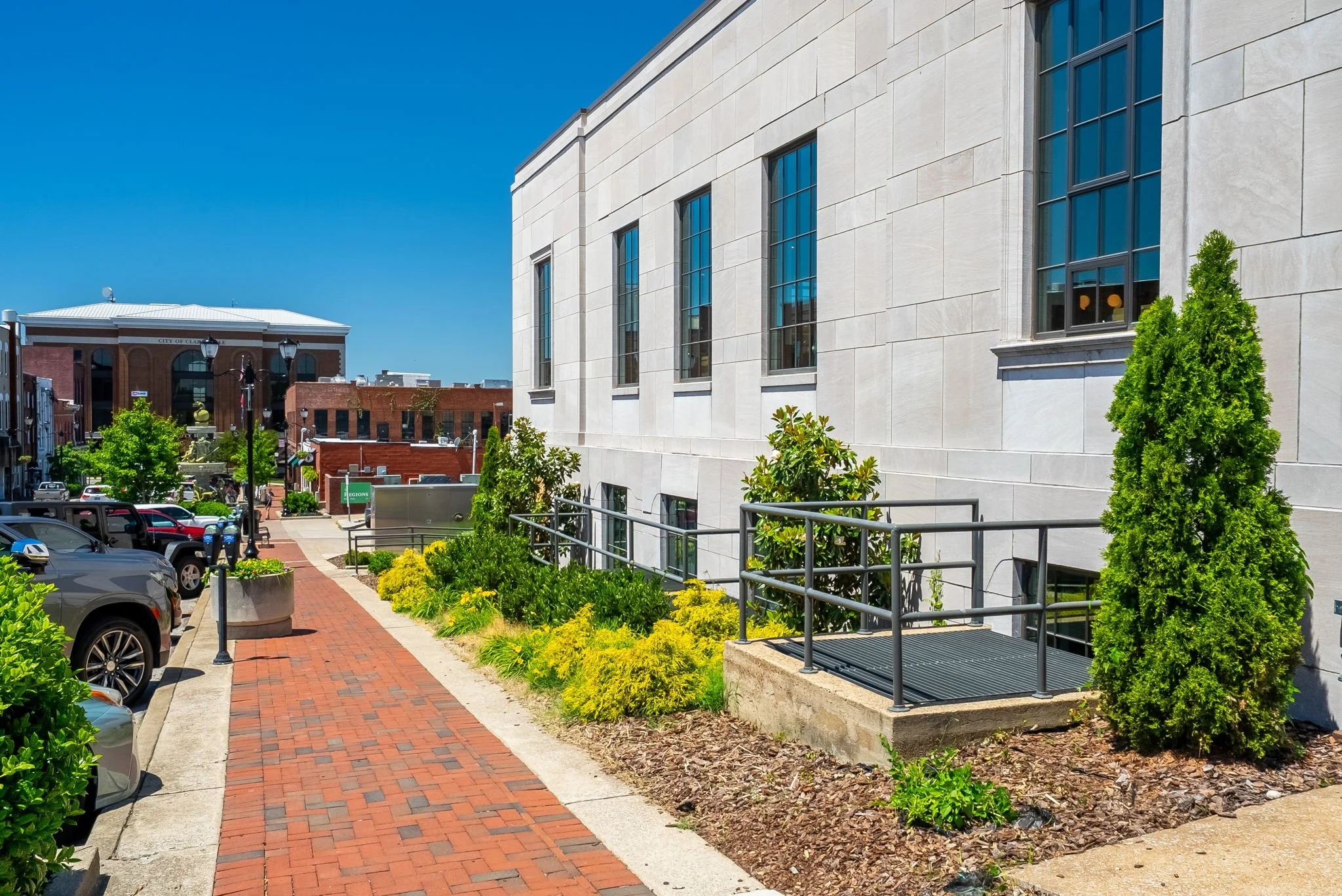 A street view with a brick sidewalk, parked cars on the left, and a large modern building with tall windows on the right. Trees and shrubs are along the sidewalk under a clear blue sky.