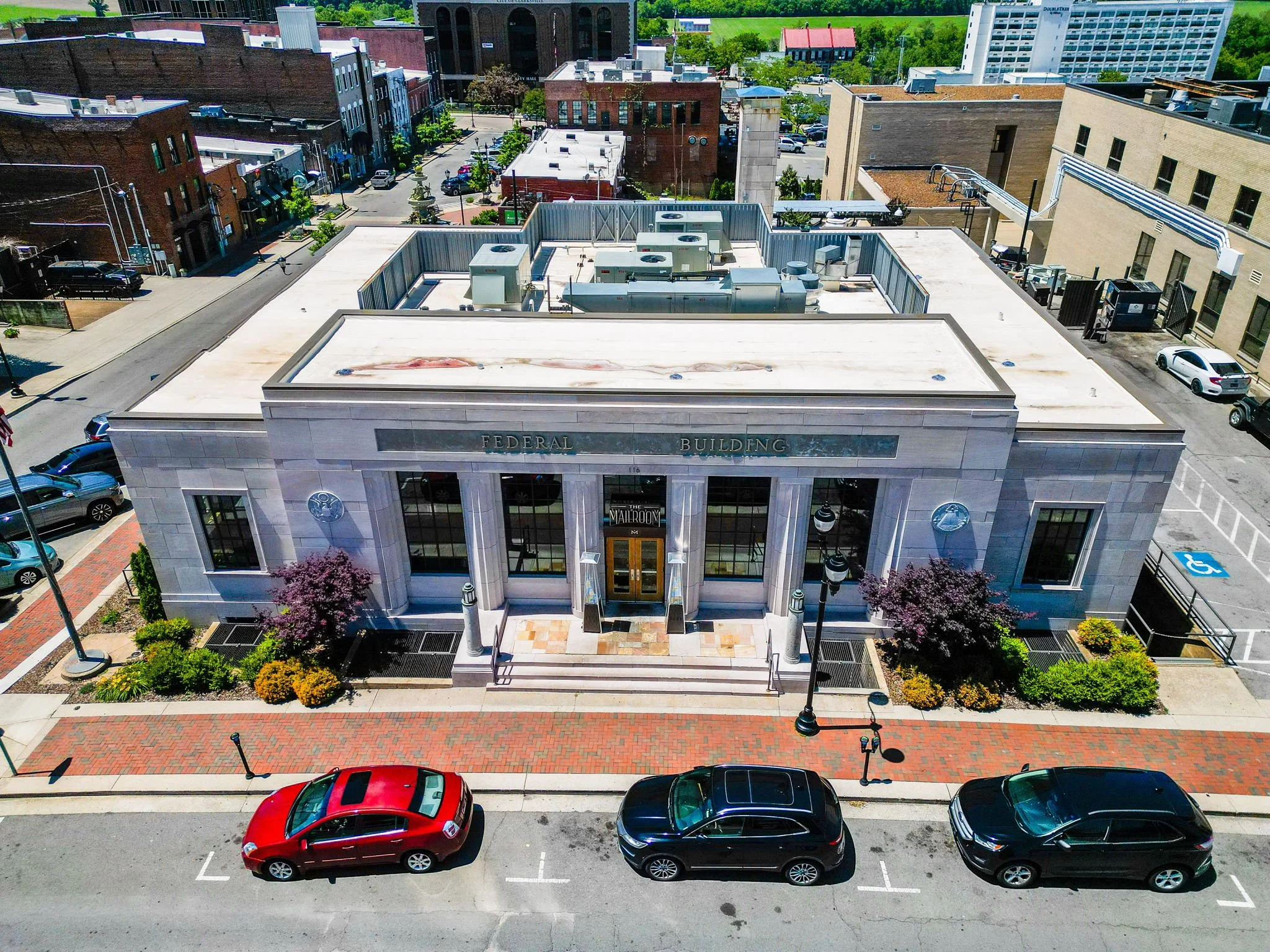 Aerial view of a city street with a building labeled 'Federal Building' on the front. Several cars are parked along the street, and small trees and plants line the sidewalks. The rooftops of nearby buildings are visible, along with a parking garage i