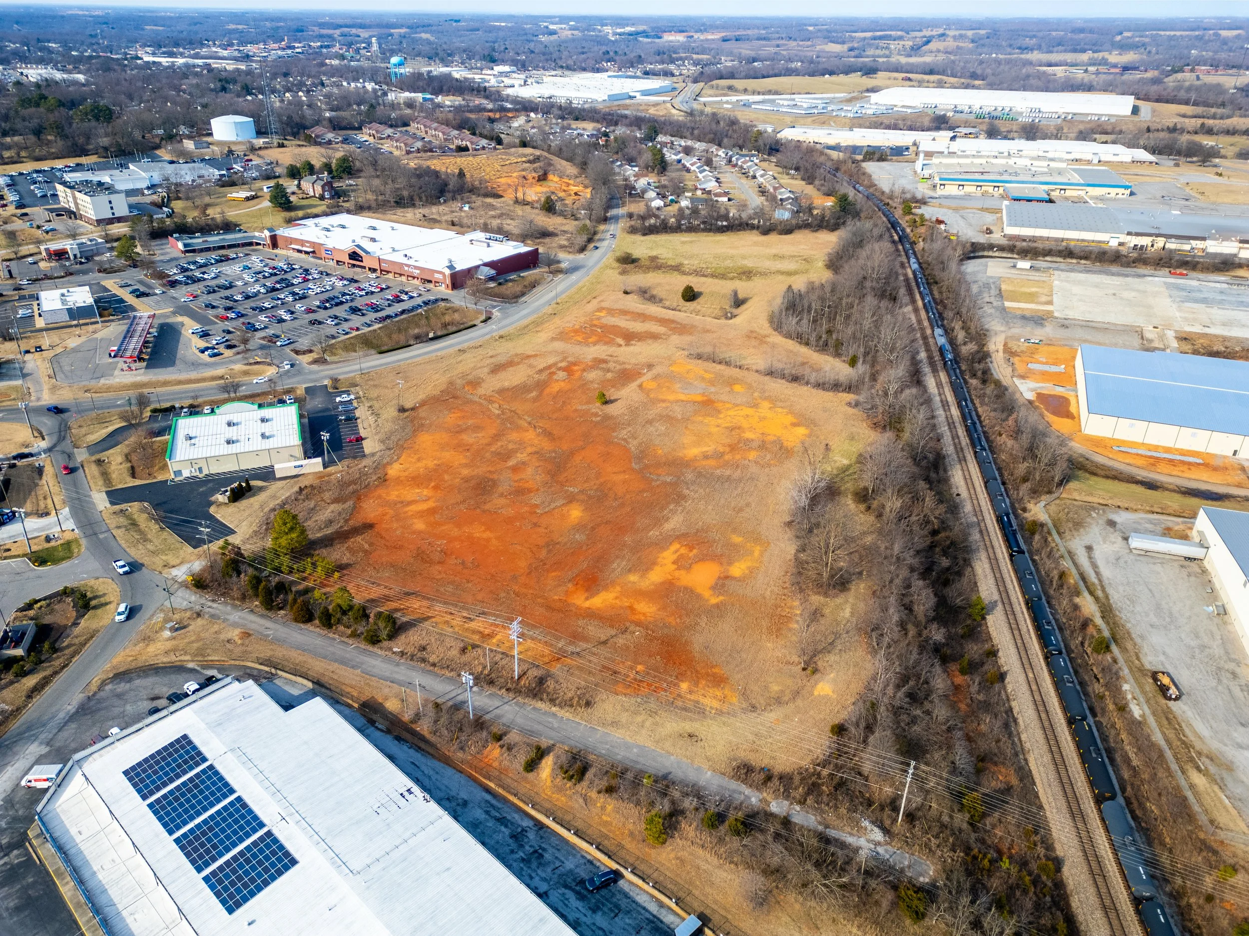 Aerial view of a commercial area with a parking lot, industrial buildings, and a train passing through a grassy and wooded area.
