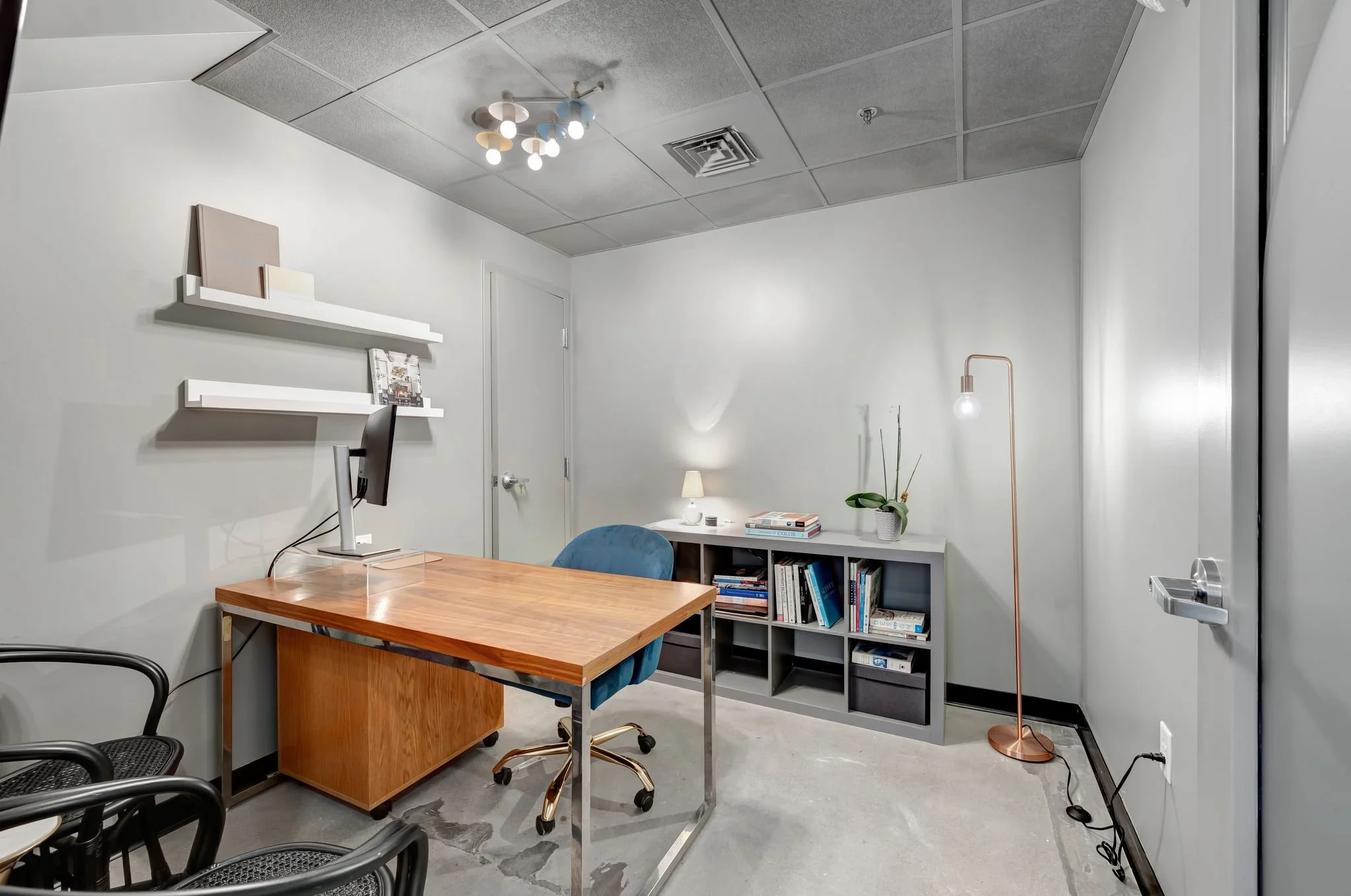 An office with a wooden desk, a computer monitor, a blue office chair, black chairs, white wall shelves, a bookshelf with books, a small lamp, a potted plant, a tall floor lamp, and a ceiling light fixture.