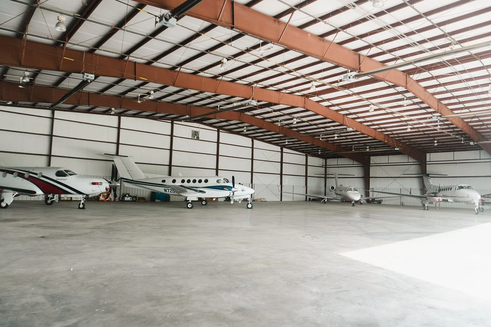 Inside a spacious airplane hangar with four private jets parked on the floor, metal beams and a high ceiling, with some equipment and supplies around.