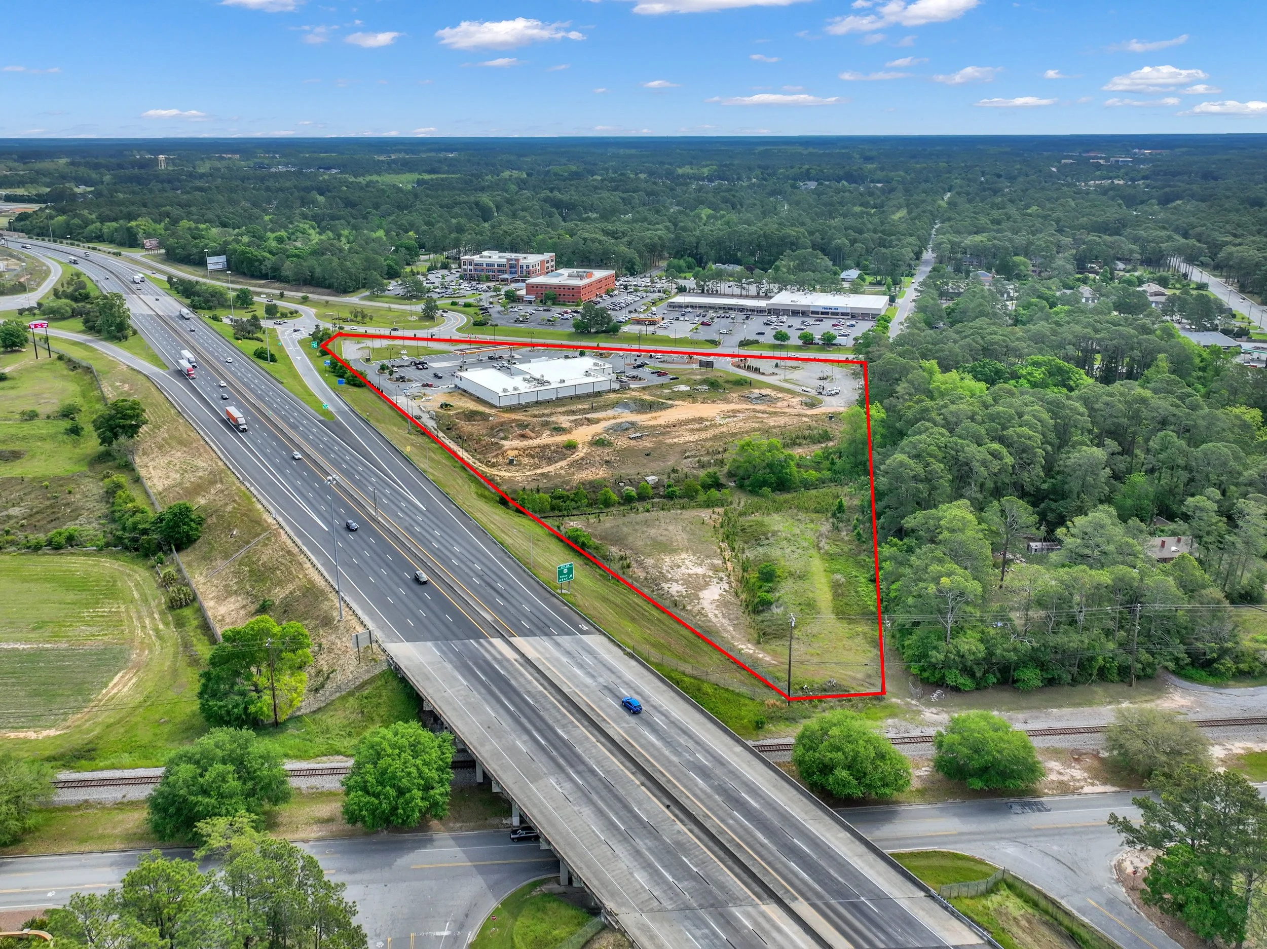 Aerial view of a construction area outlined in red near a highway, with a commercial shopping area in the background and green wooded areas surrounding the site.