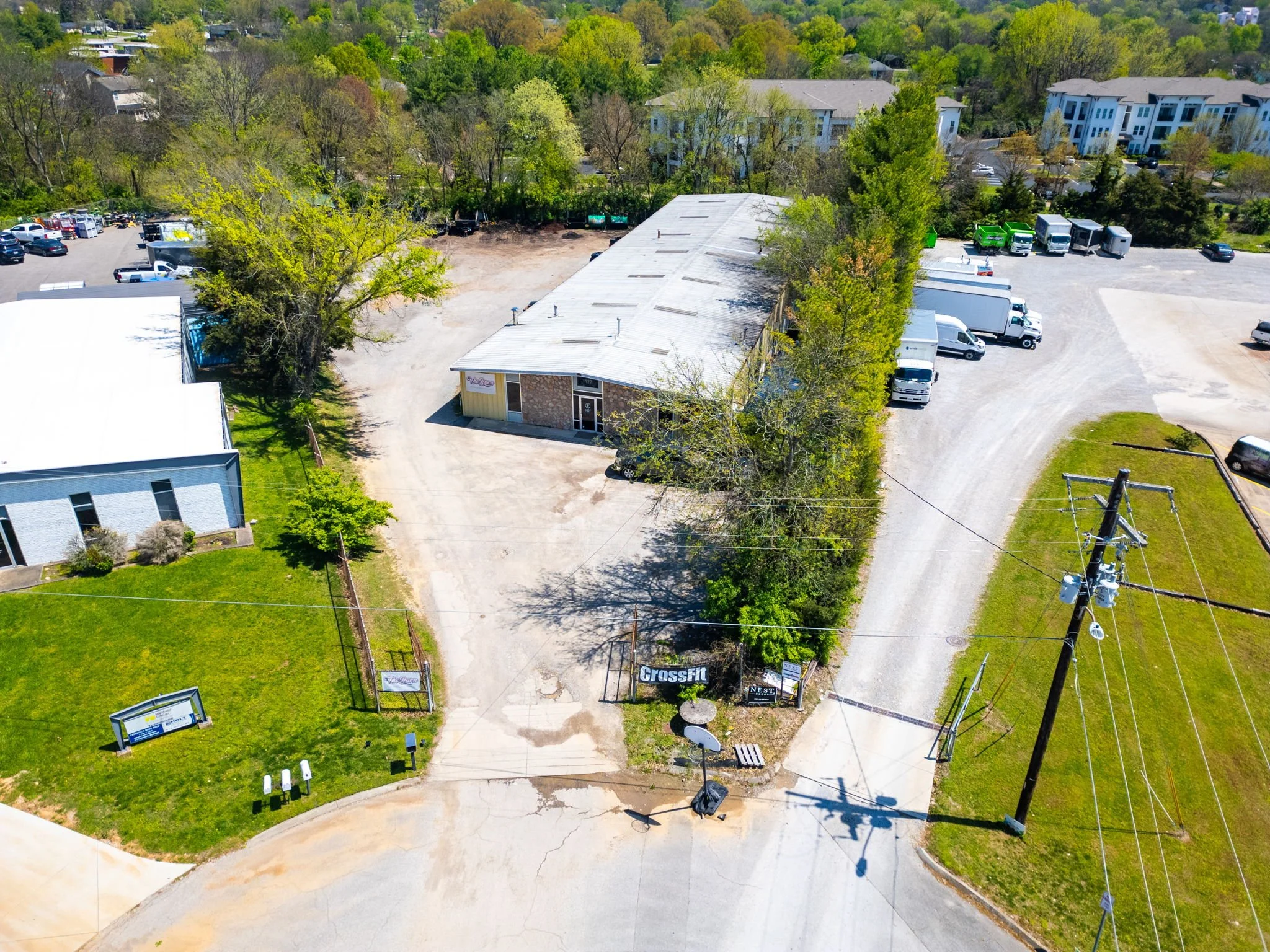 An aerial view of a commercial building with a white roof and a parking lot, surrounded by trees and other buildings. There are several trucks parked along the side of the building and a road leading into the property. Signage for Crossfit is visible