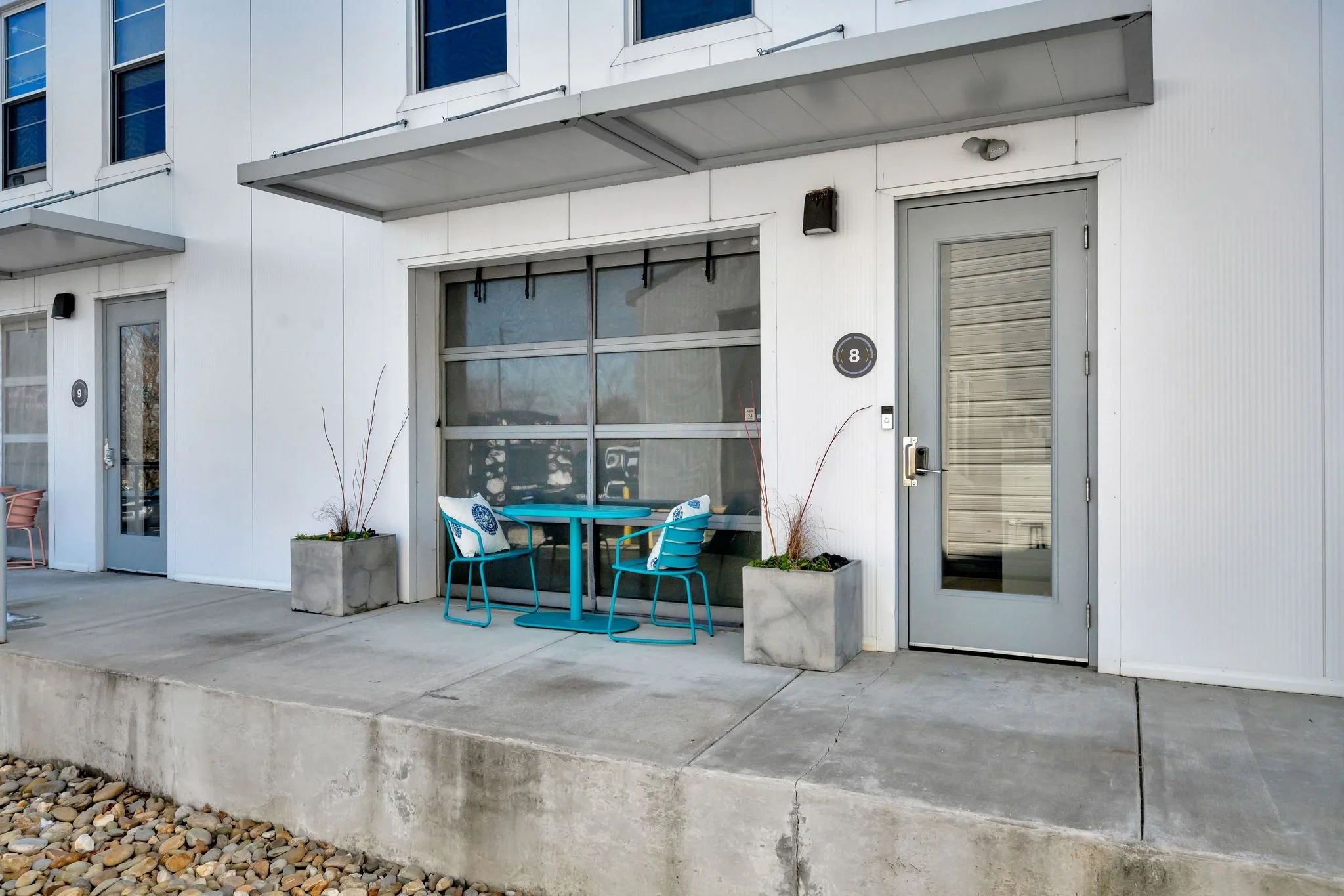 Modern apartment building with outdoor seating area featuring two blue chairs and a round blue table, in front of large glass window and door, with concrete planters and stone ground cover.