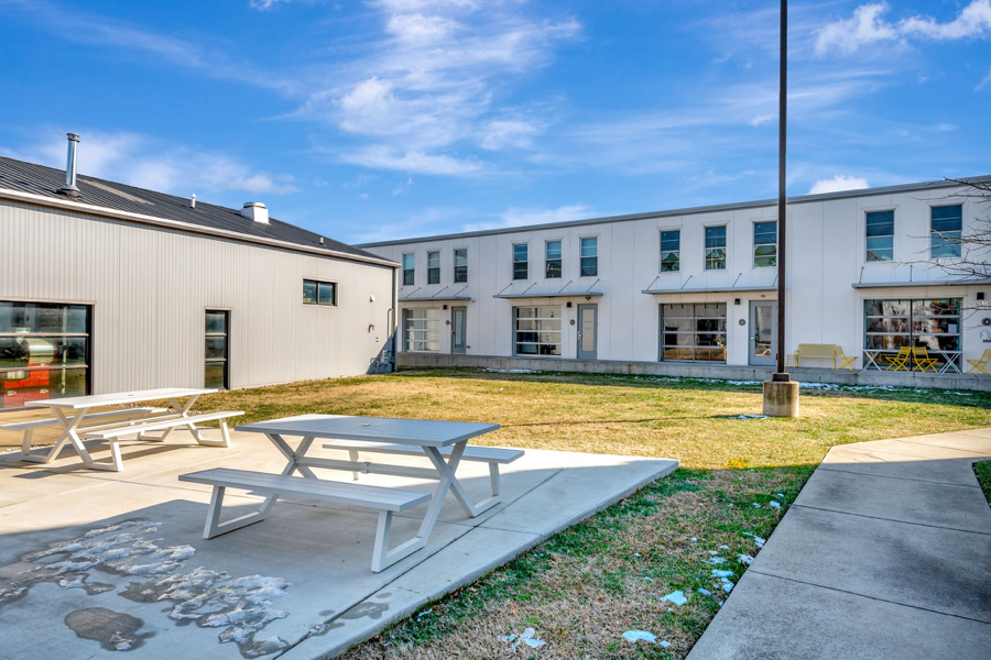 Outdoor area with concrete patio, picnic tables, grassy lawn, apartment building, and blue sky.