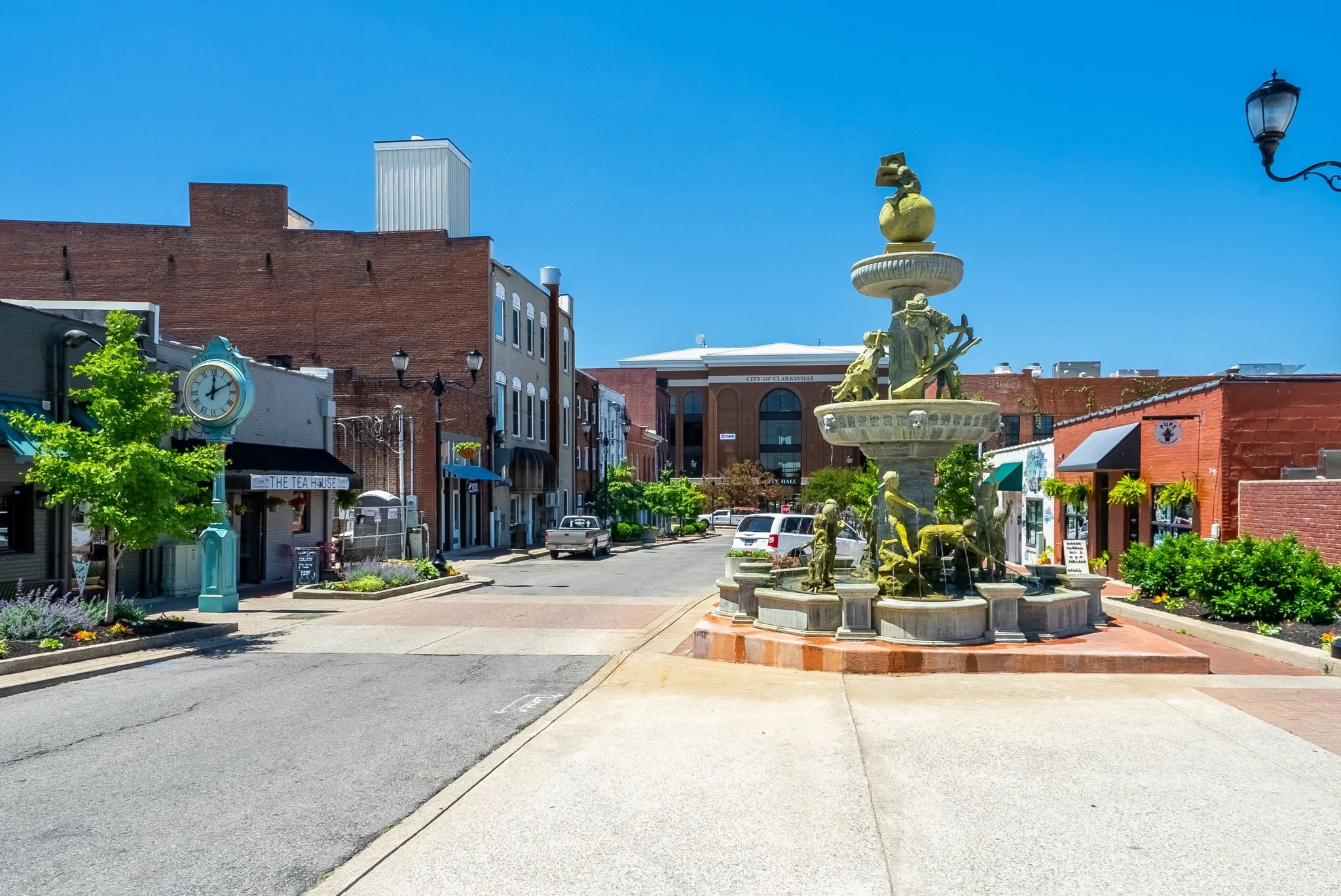 A small downtown area with shops and businesses, including a tea house and a clock, with trees and a fountain featuring bronze statues in the center. The buildings are made of brick and the sky is clear and blue.