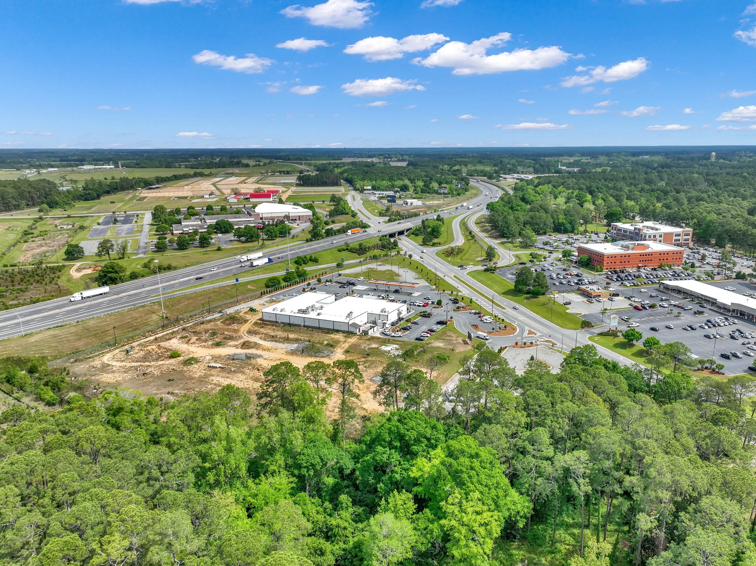 Aerial view of a commercial area and highway surrounded by trees and green landscape on a sunny day with some clouds in the sky.