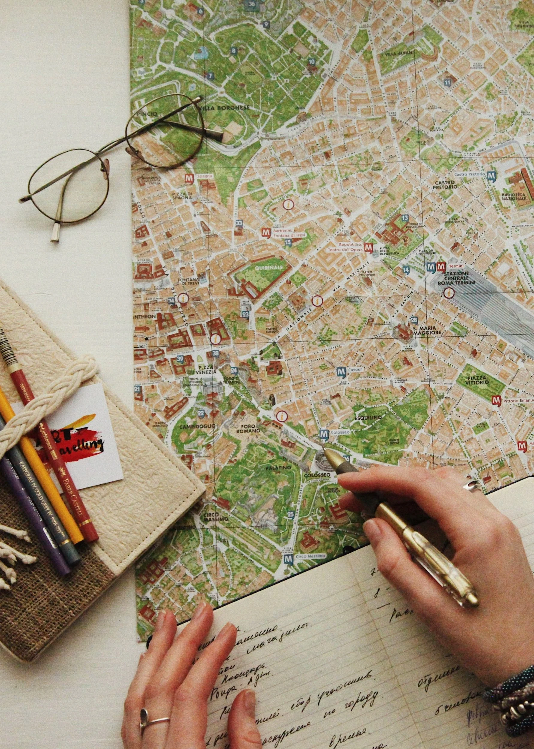 Person pointing at a city map with a pen, surrounded by glasses, a notebook, and colored pencils, planning a trip.
