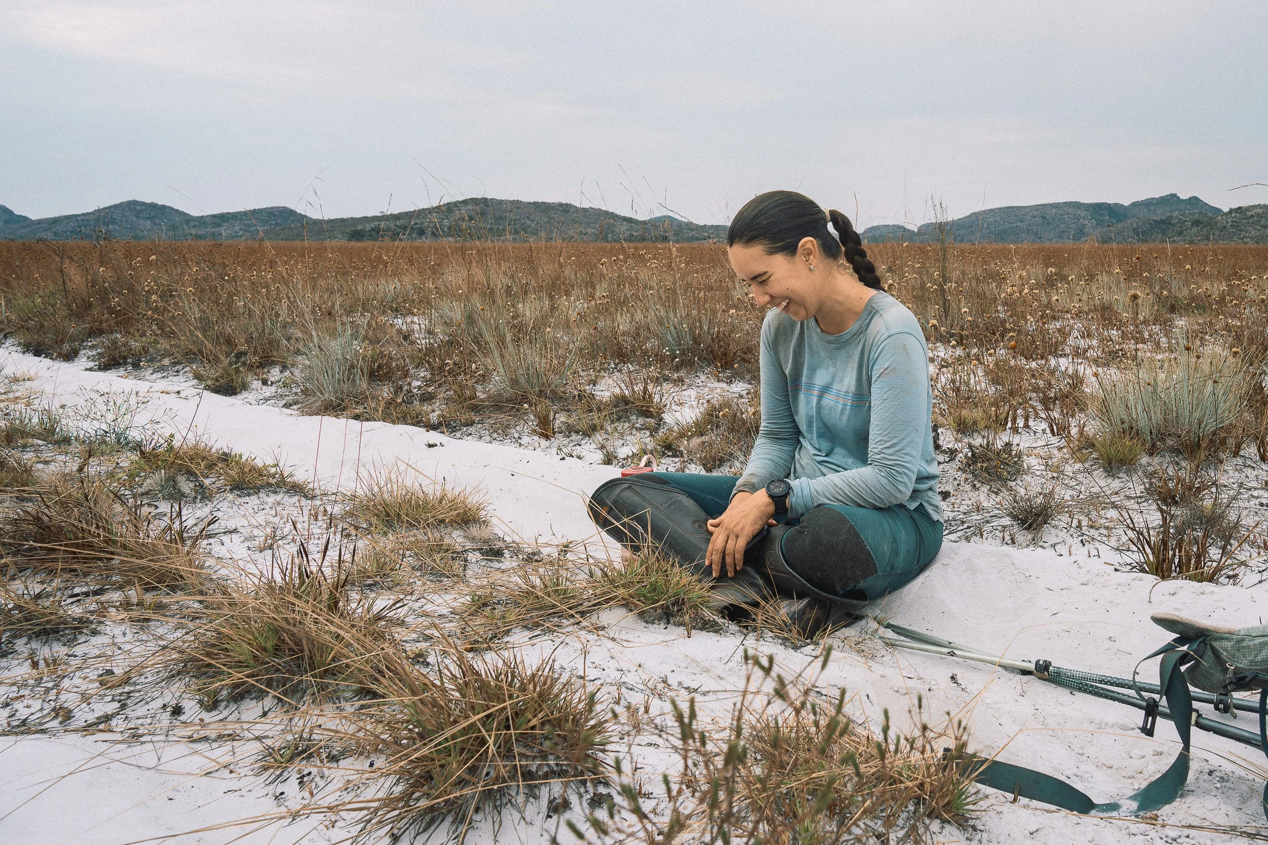 Elisa sits smiling on white sand among tall grasses and flowers during a trek across Campos Tristes in Sempre Vivas National Park