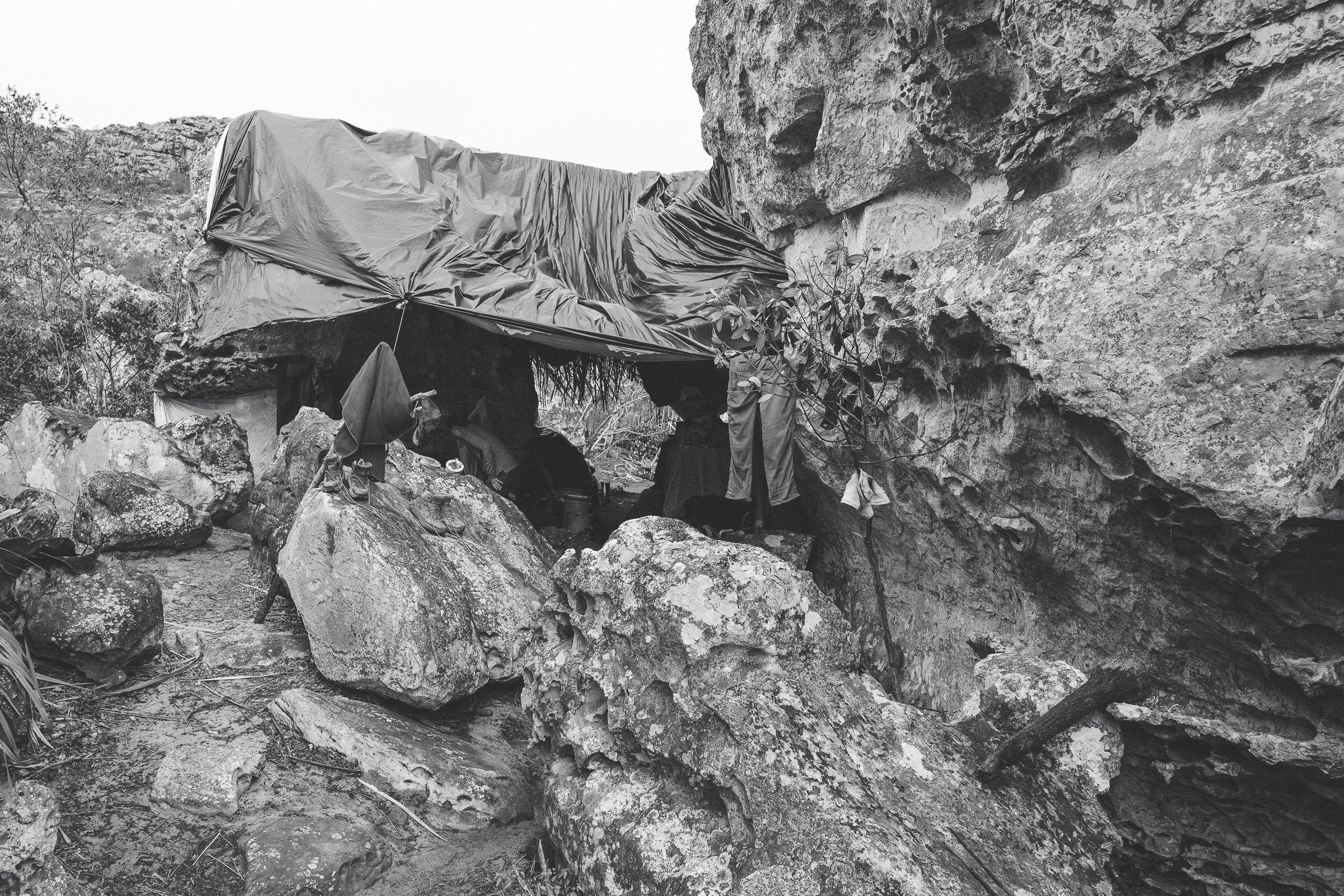 Black-and-white view of a makeshift tarp roof stretched between boulders at Leandro’s rock shelter in Campos Tristes, Sempre Vivas National Park