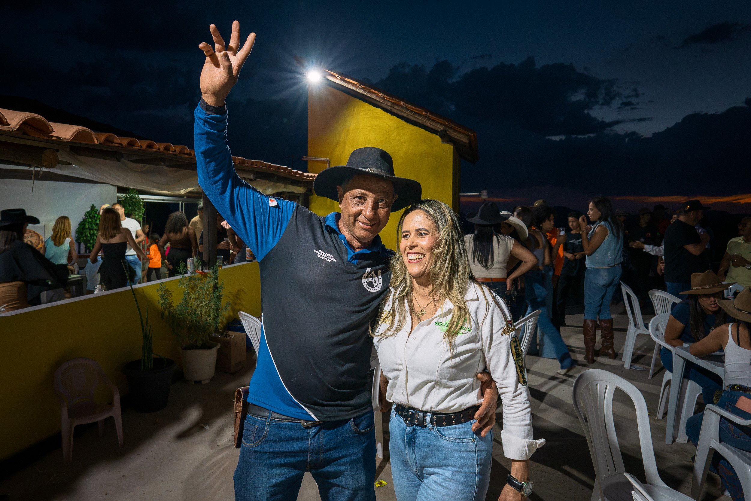 Pê, wearing a black cowboy hat, raises his hand beside his wife Nica during Branquinho’s 50th birthday party near Guinda, Diamantina.