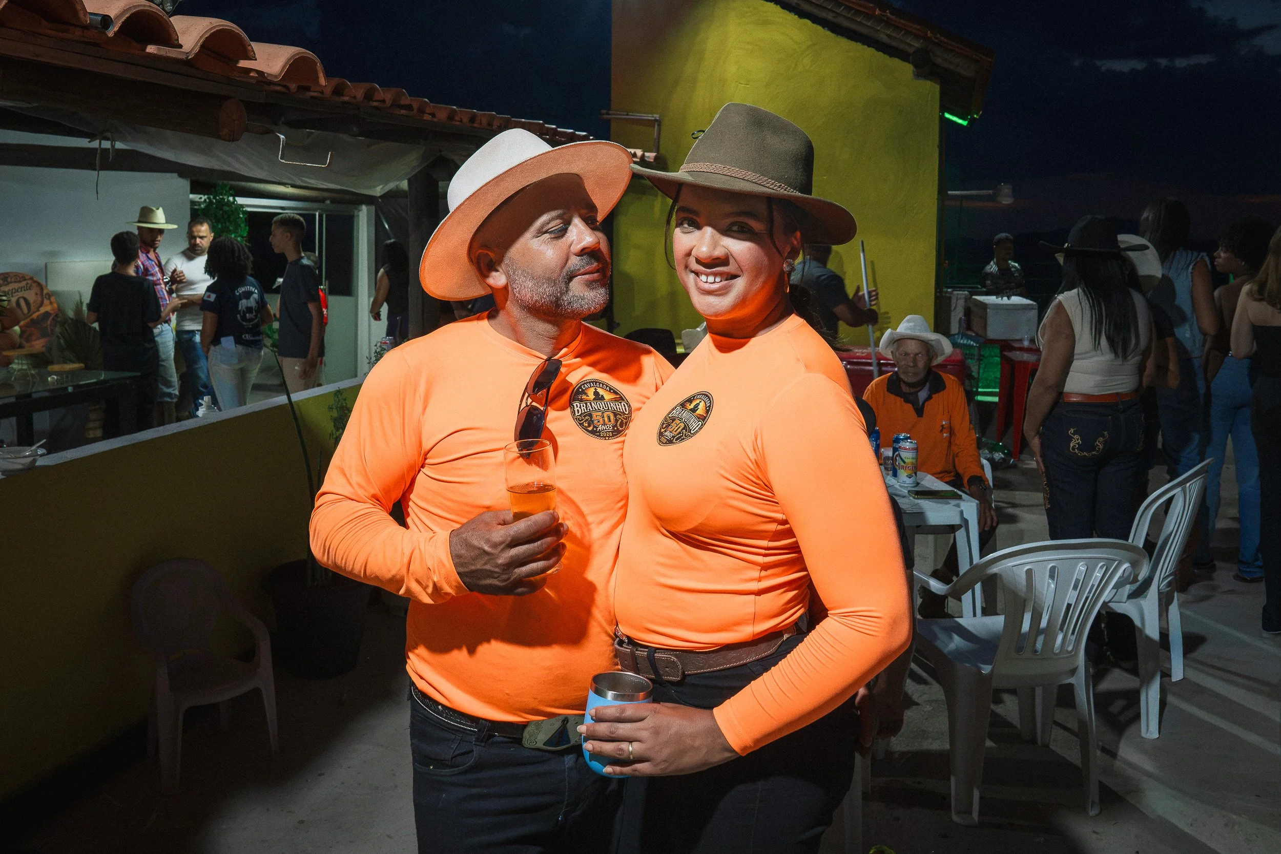 Branquinho, in a white cowboy hat and bright orange shirt, stands with his wife Ana Cláudia at his 50th birthday party near Guinda, Diamantina.