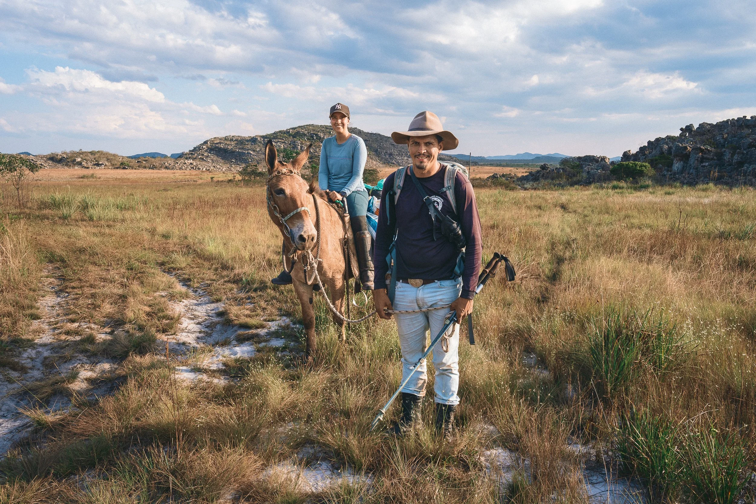 Elisa sits on Baiana beside Guim, who holds the reins on a grassy trail between rocky hills in the Serra do Espinhaço.