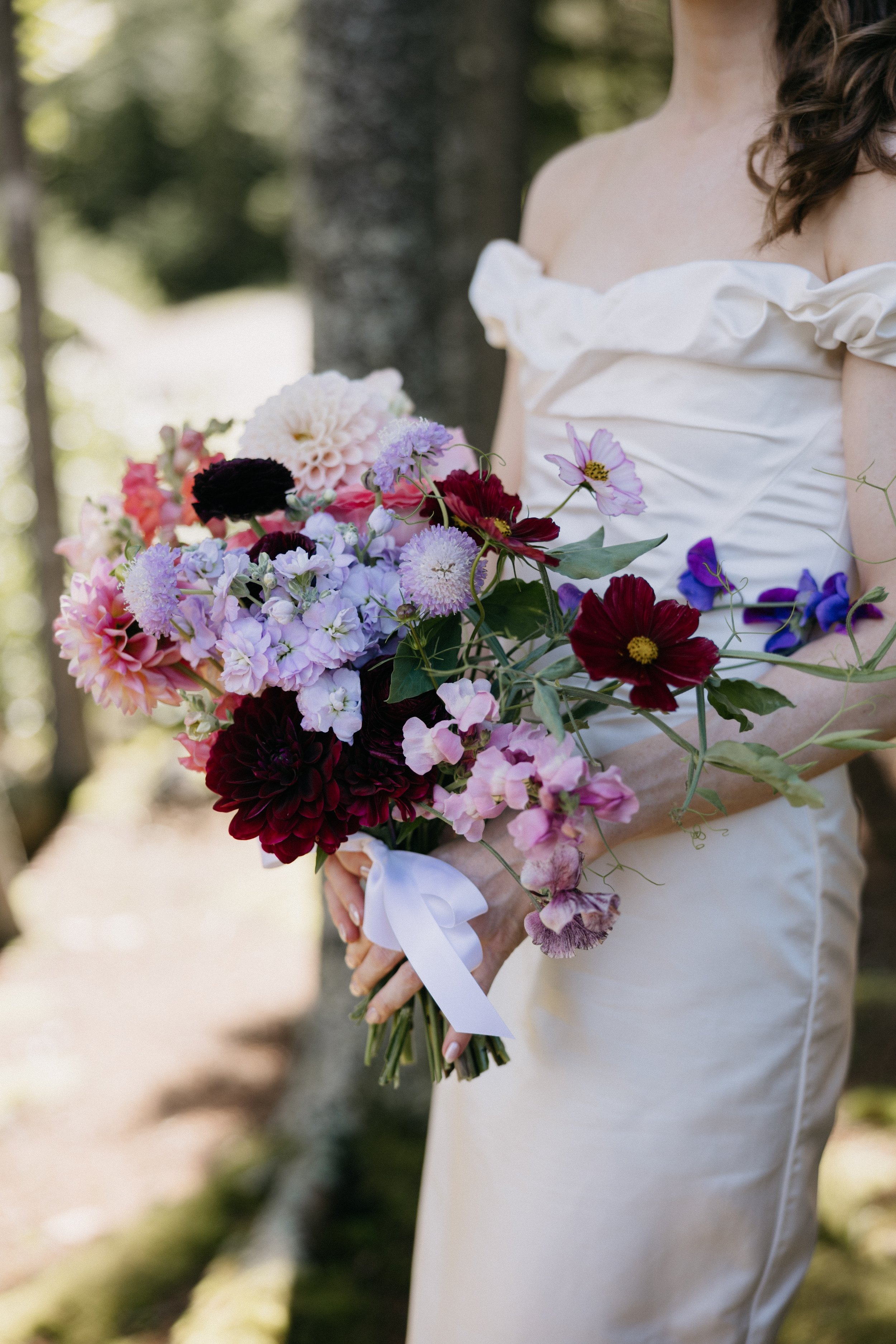 A woman in a white off-shoulder dress holding a colorful bouquet of flowers with purple, pink, red, and white blooms, with green foliage, tied with a white ribbon in an outdoor setting with trees and sunlight.