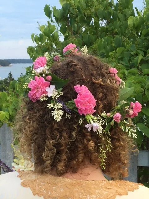 Person with curly hair wearing a floral crown of pink, white, and purple flowers, outdoors with green foliage and water in the background.