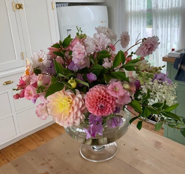 A large glass bowl filled with pink, purple, and white flowers on a wooden table in a bright room.