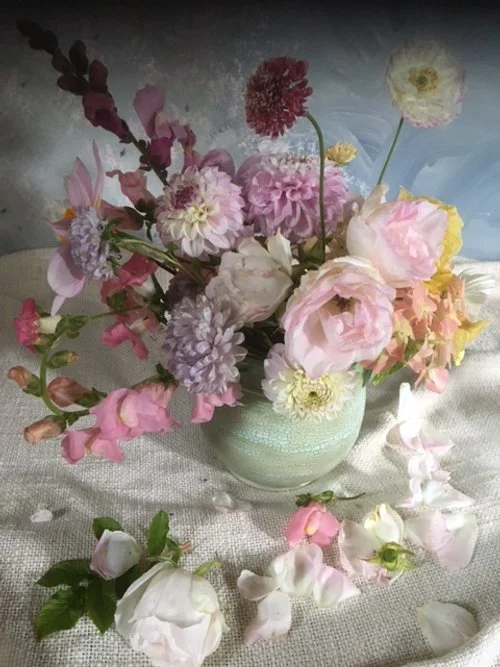 A bouquet of various pink, white, and purple flowers in a light green vase, with some flowers and petals scattered on a textured surface.