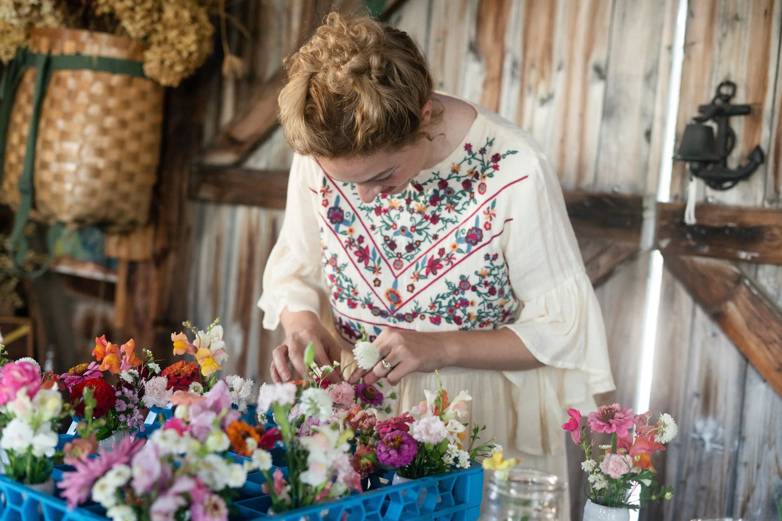 Woman in a white embroidered blouse arranging colorful flowers in a basket inside a rustic wooden barn.