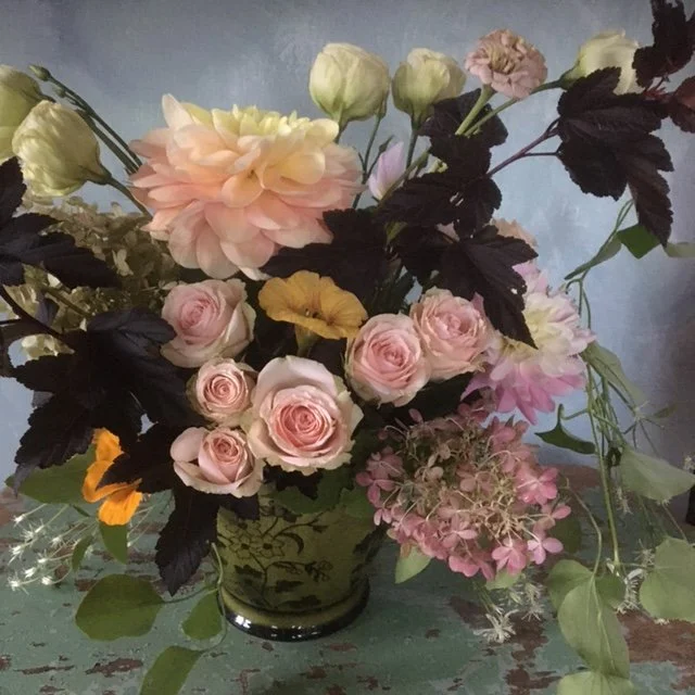 A floral arrangement in a dark vase featuring pink roses, peach dahlias, white lisianthus, and pink hydrangeas alongside dark green and black leaves.