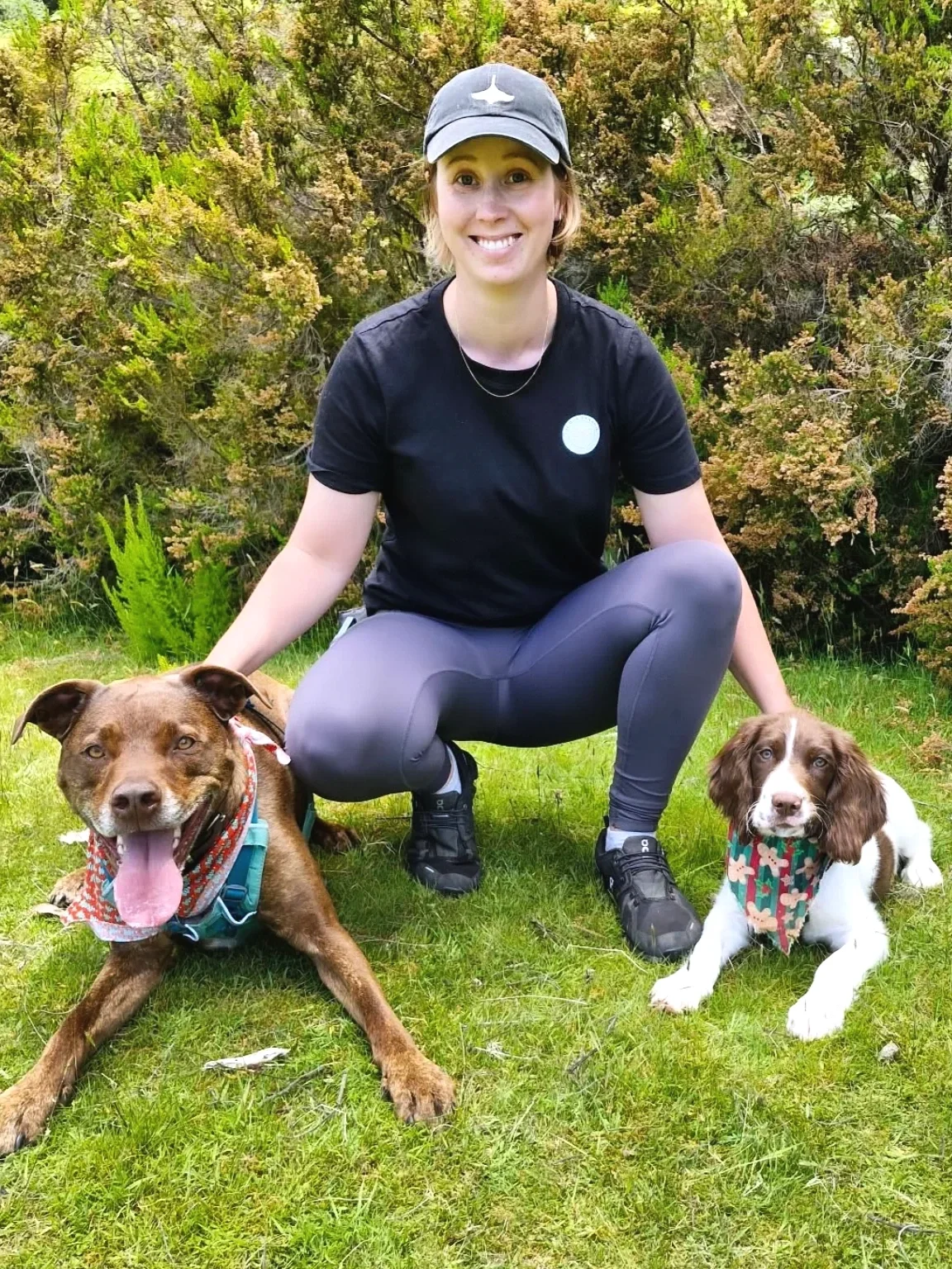 A woman crouching on grass outdoors with two dogs, surrounded by green shrubbery.