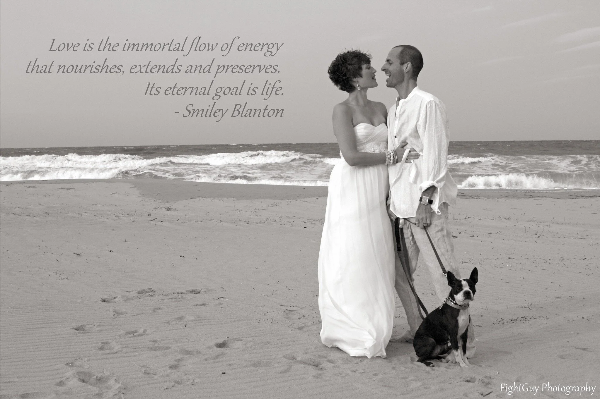 A couple stands on the beach at their wedding, with their Boston Terrier sitting at their feet