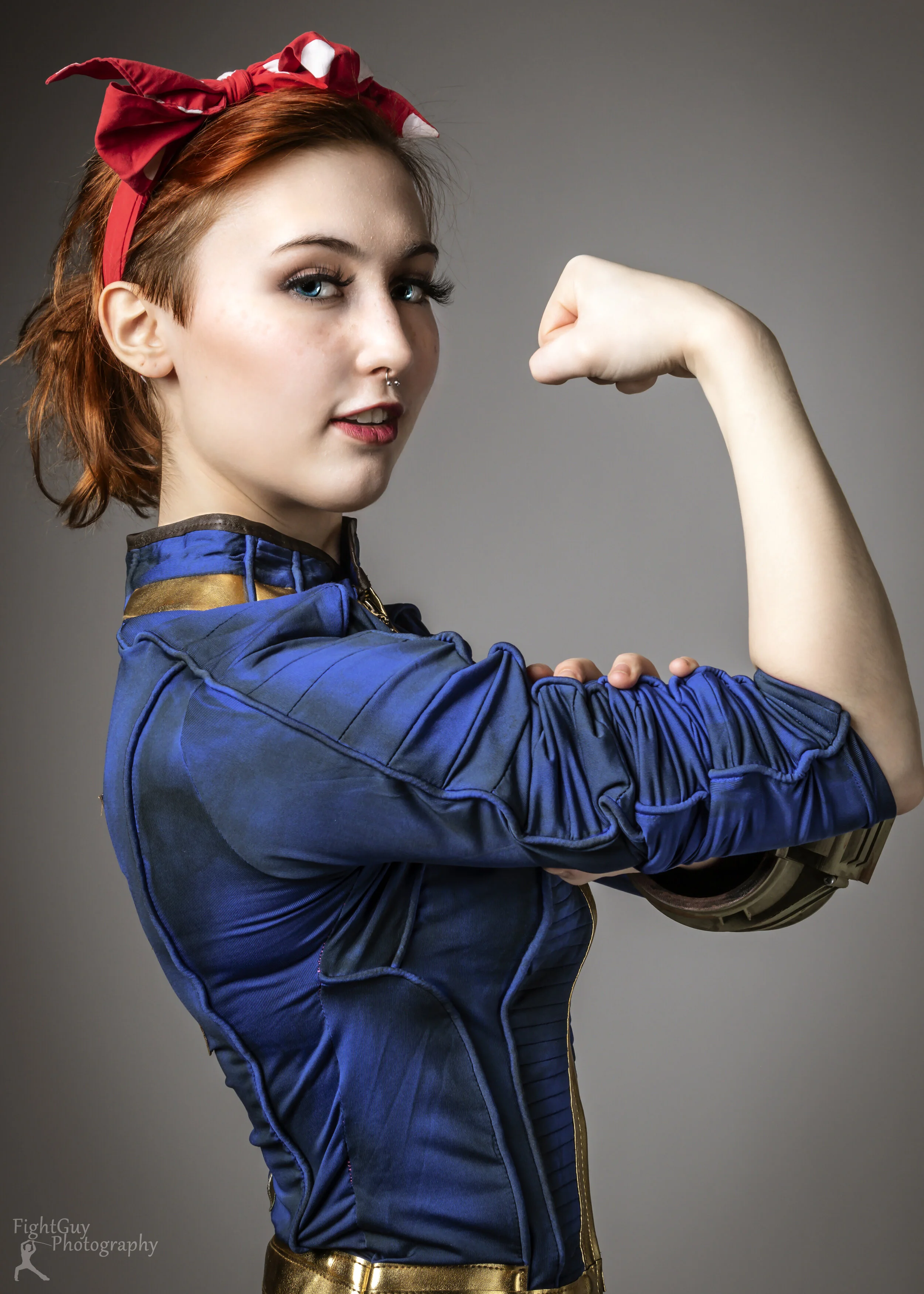 A redhead in a blue jacket, with a red bandanna in her hair, strikes a Rosie the Riveter Pose.