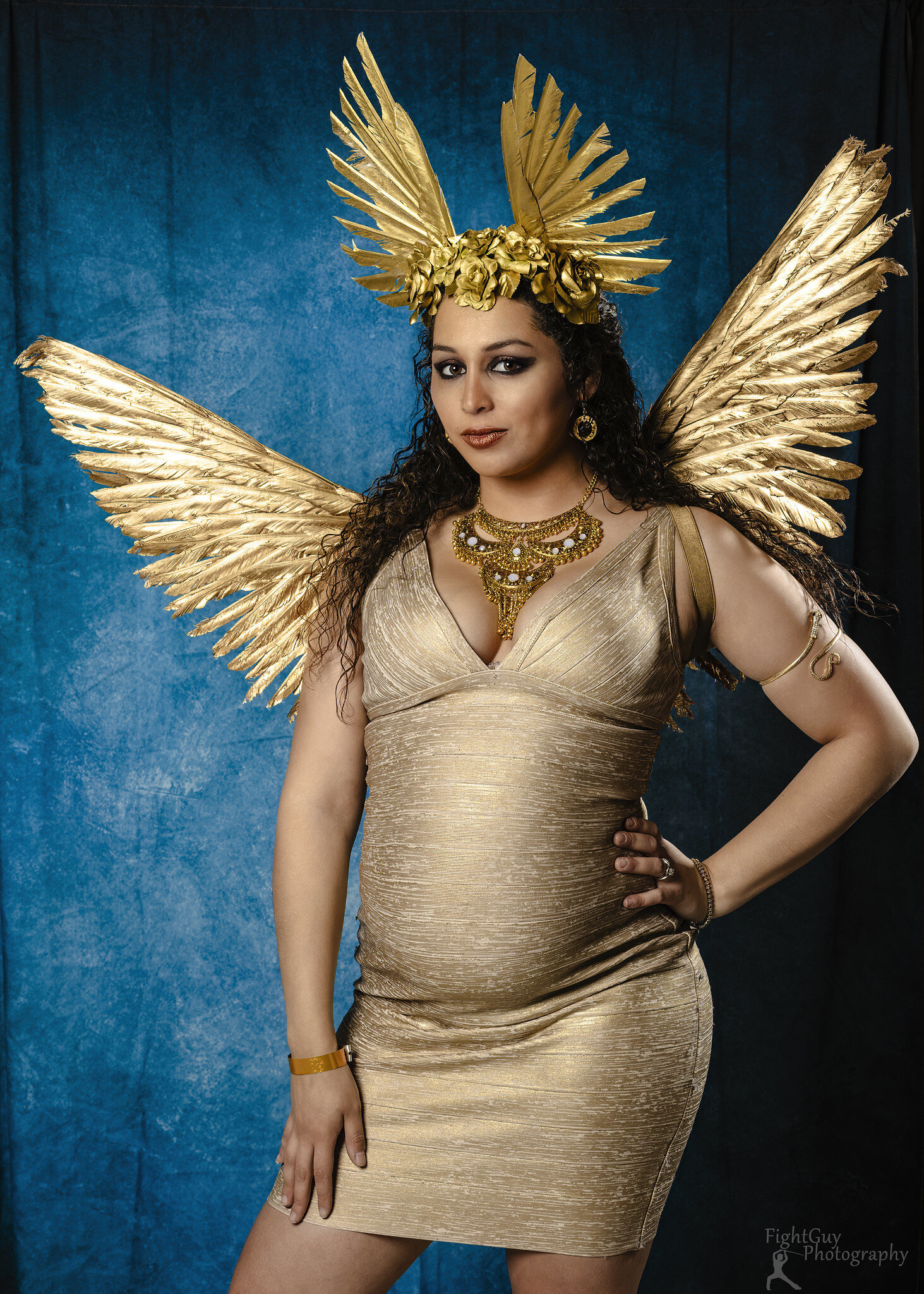 Woman wearing a gold dress, ornate gold headpiece with wings, wing-shaped accessories on her shoulders, gold jewelry, standing against a blue background.
