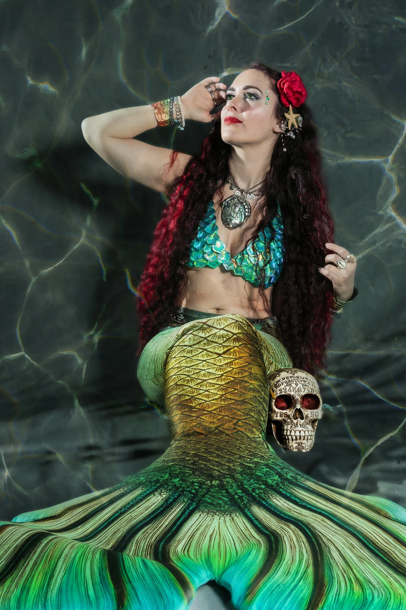 A woman dressed as a mermaid with long red hair, wearing a shiny blue scale top and green tail, floating in water with a skull beside her.