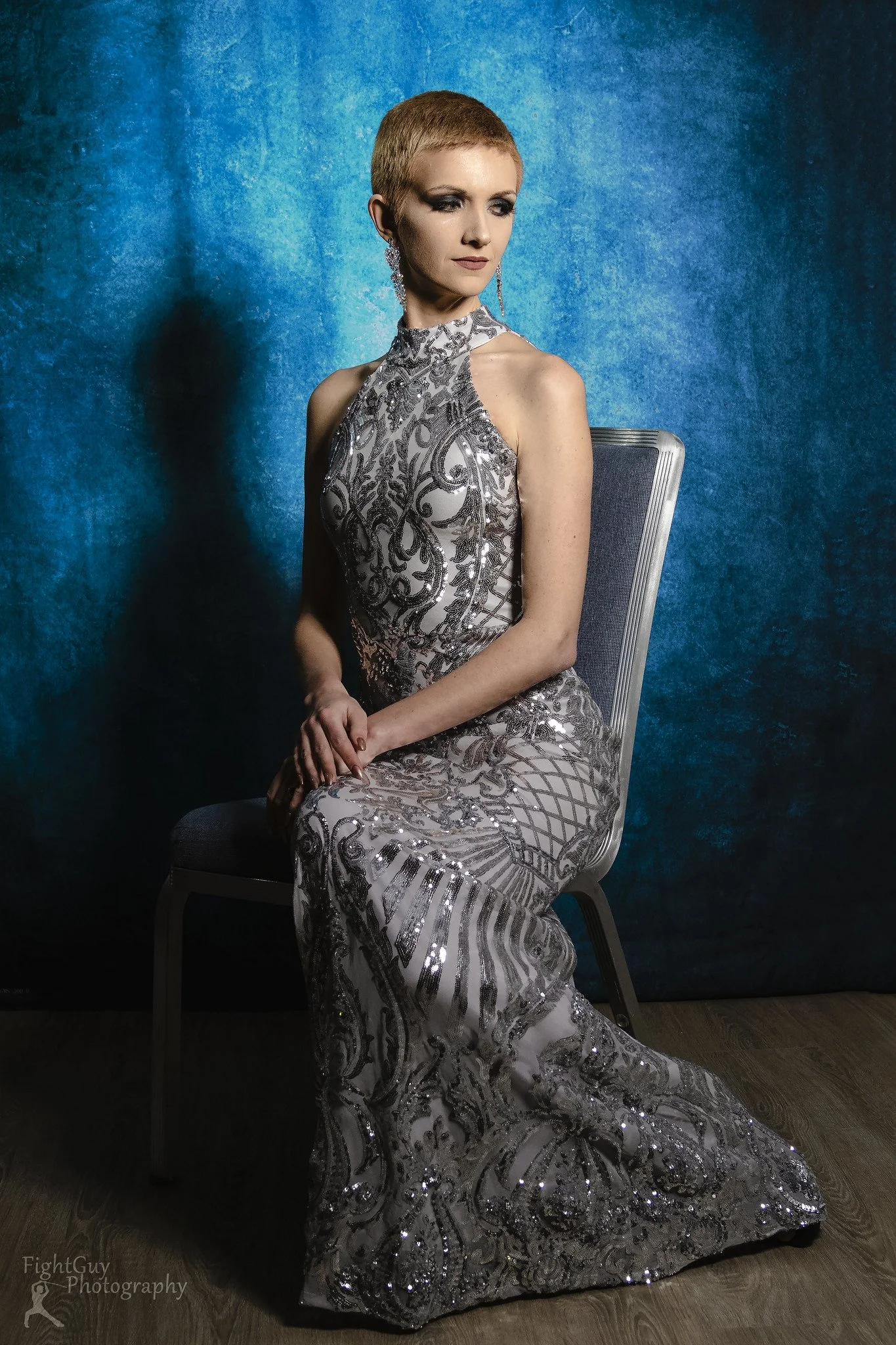 A woman with short blonde hair sitting on a chair, wearing an elegant silver and black patterned evening gown, with a blue textured background.