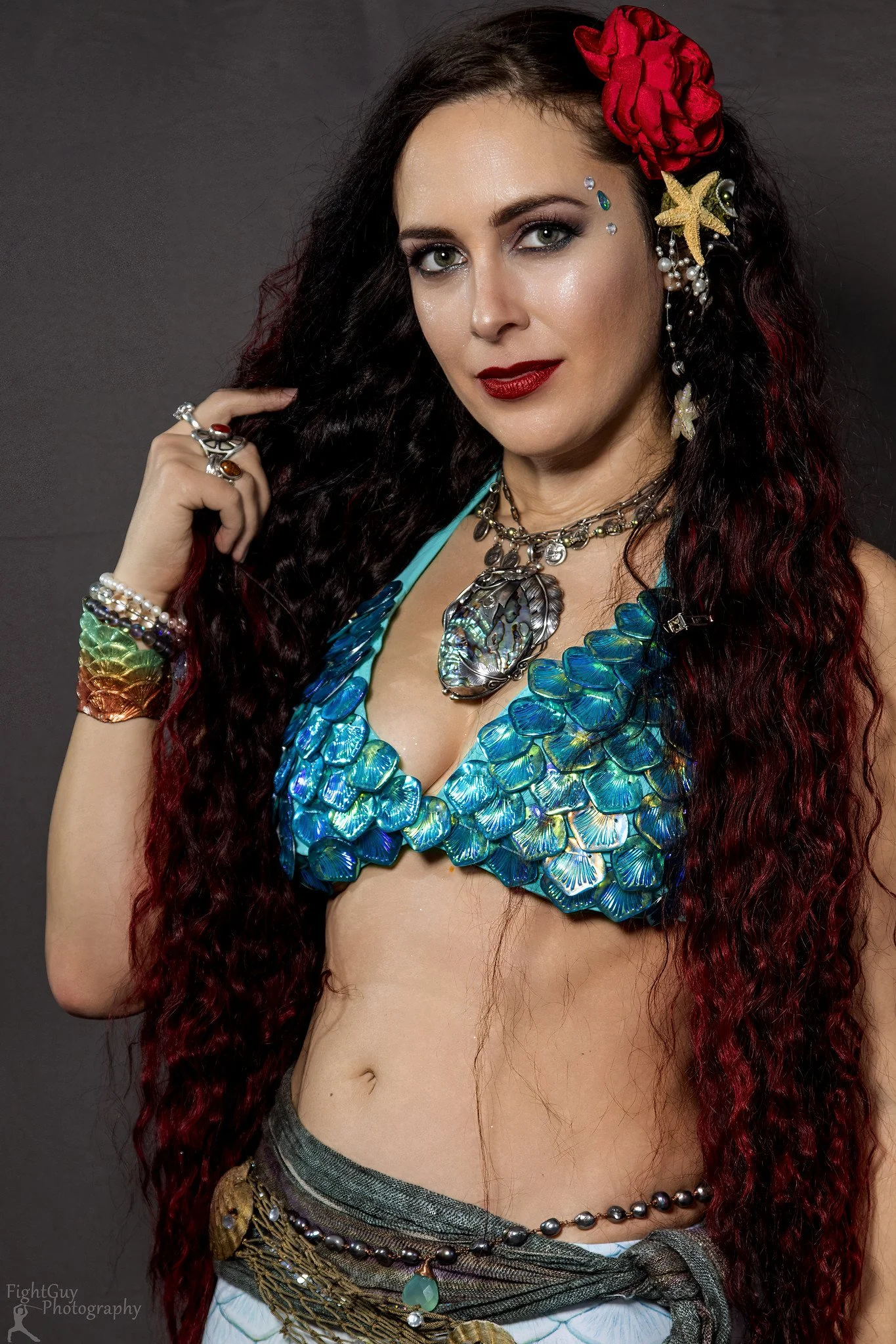Woman with long dark curly hair wearing colorful jewelry, red flower hair accessory, decorated face, and a blue shell top, posing against a dark background.