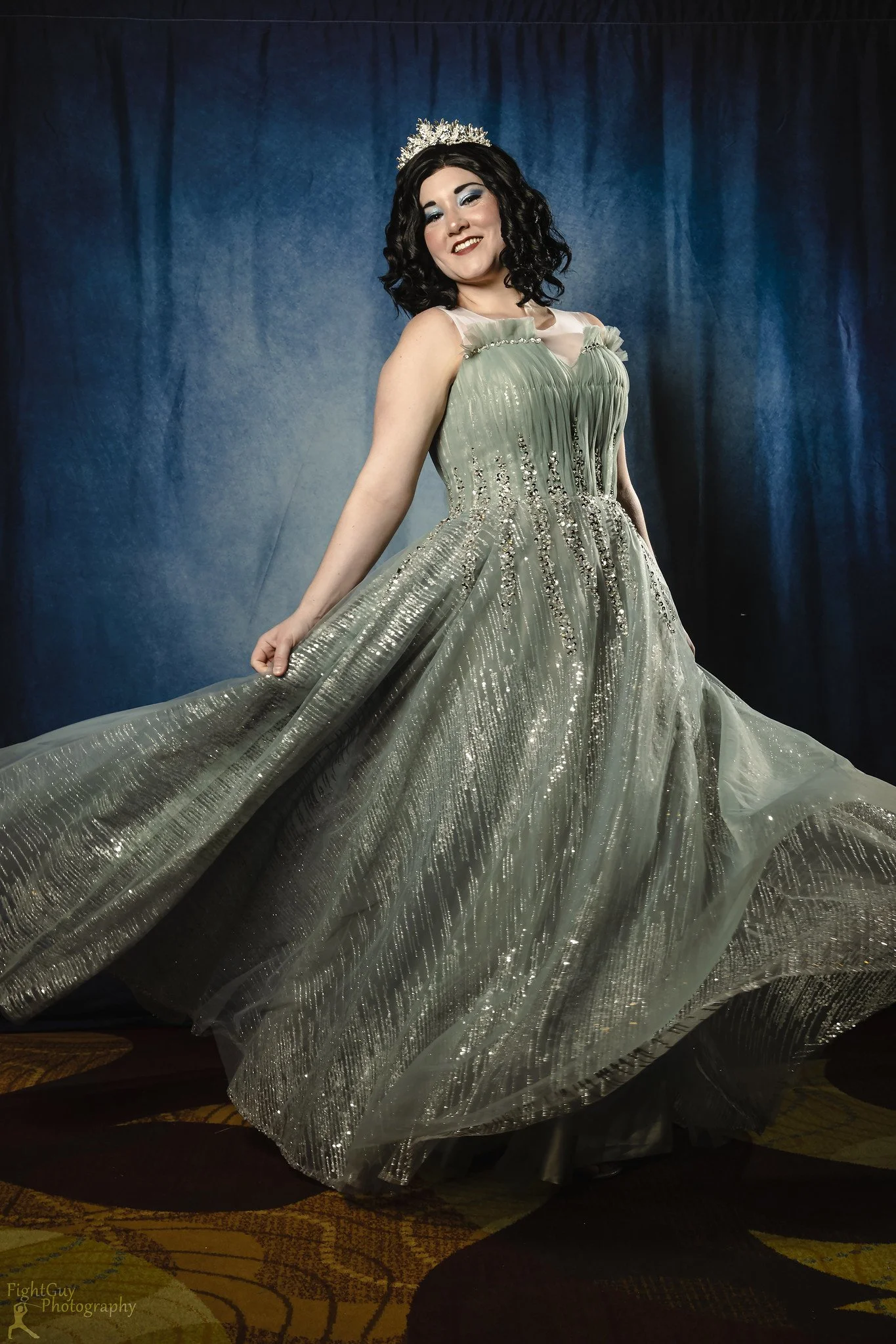 Woman wearing a silver ball gown and a tiara, standing in front of a dark blue curtain.