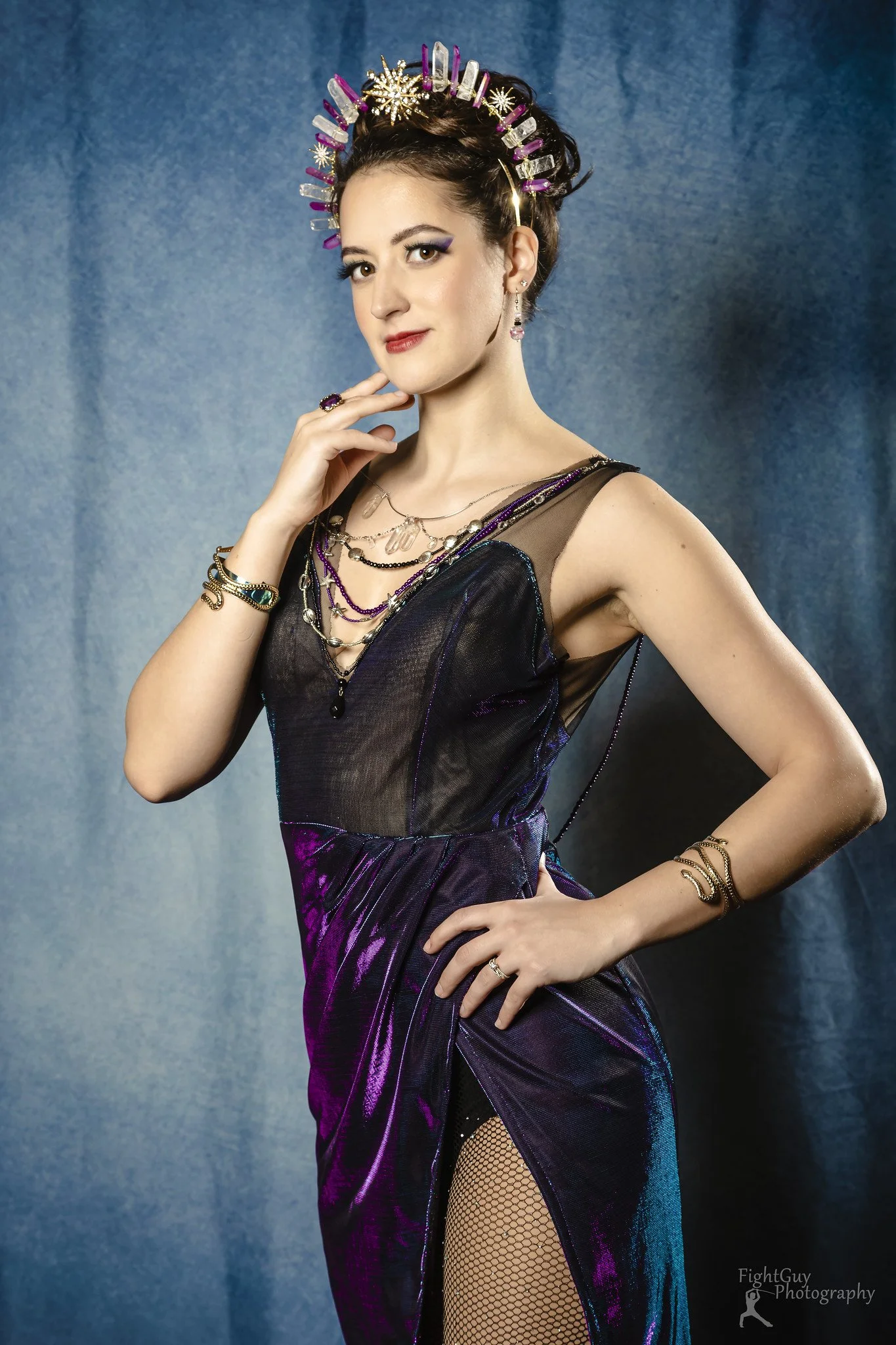 A woman in a purple and black evening gown with a slit, wearing jewelry and a decorative headpiece, standing against a blue backdrop.