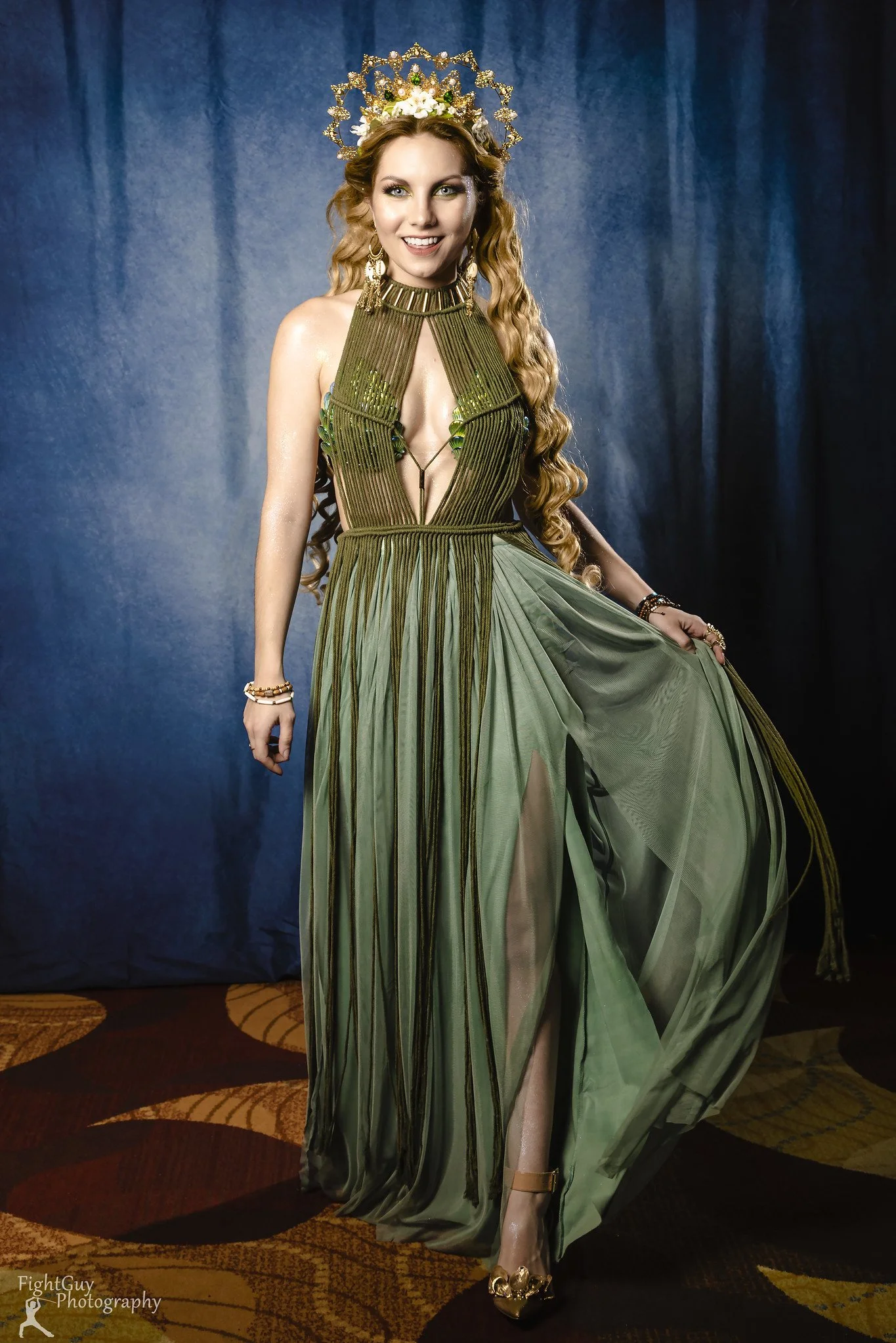 A woman wearing an elaborate green dress with a deep neckline, sheer skirt, and gold accessories, including a crown, standing on a patterned carpet against a blue backdrop.