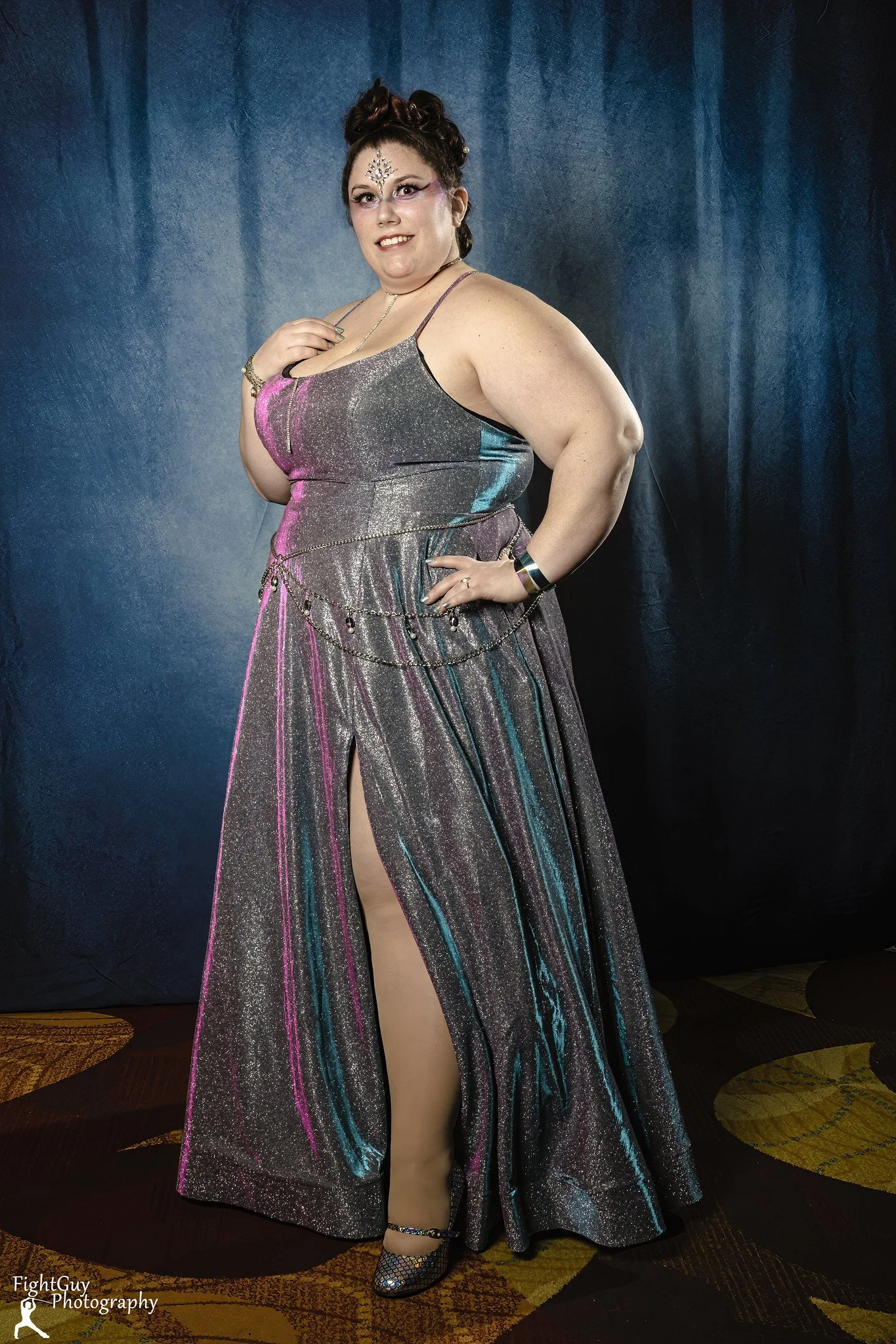 A woman in a shiny, colorful dress with a high slit, standing against a dark backdrop, smiling and posing confidently.