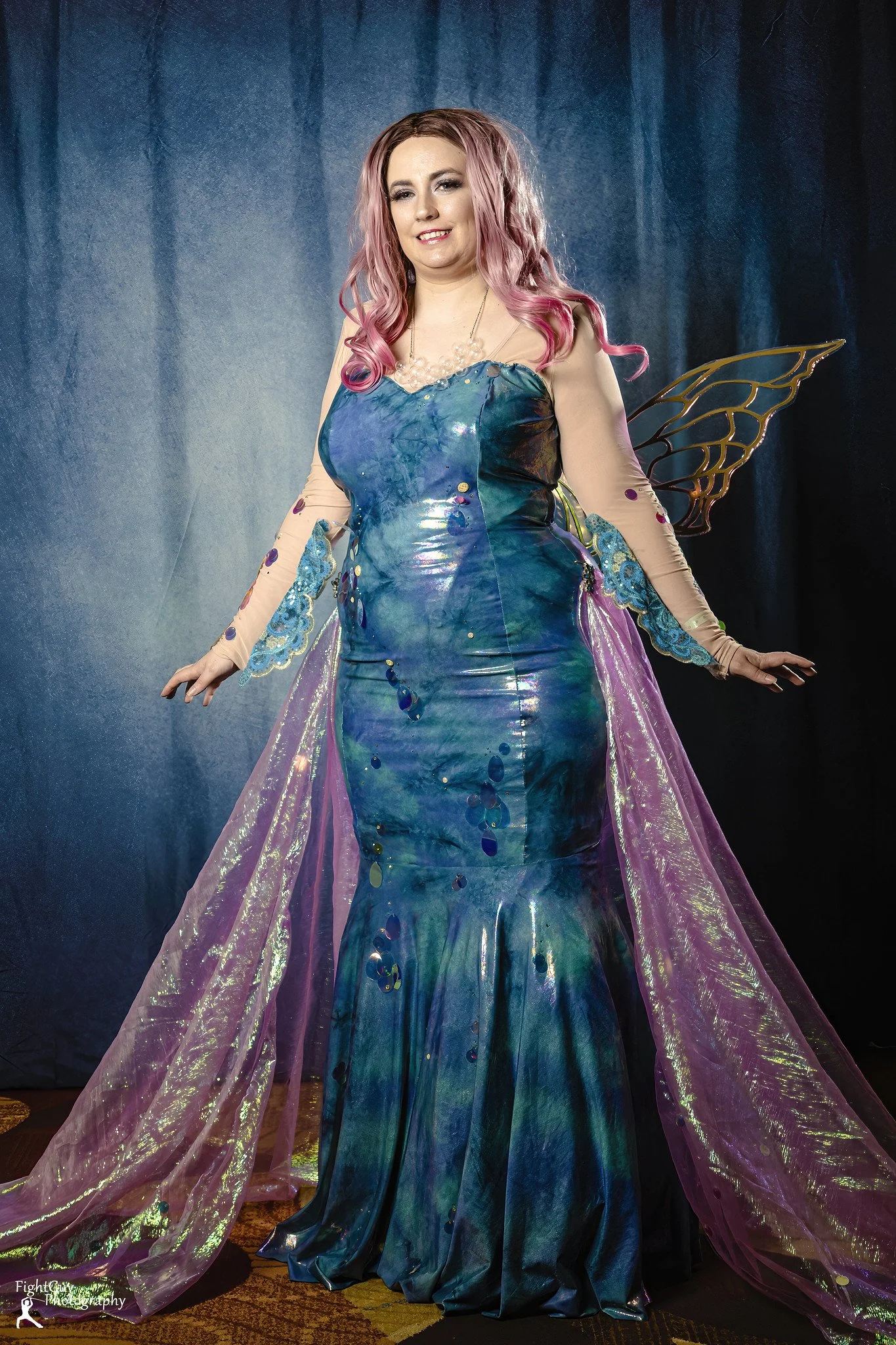 Woman in a mermaid-style dress with iridescent blue-green fabric, fairy wings, pink curly hair, and light makeup, standing in front of a dark blue backdrop.