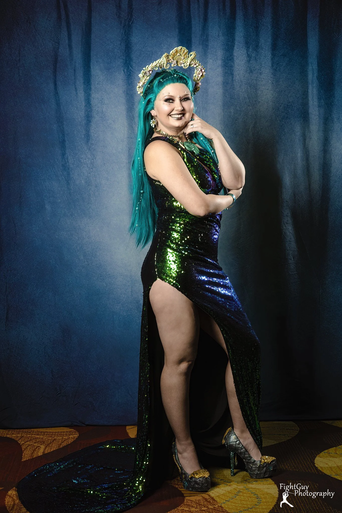 A woman with long teal hair wearing a shiny, black and green, high-slit dress and elaborate accessories, standing against a dark blue backdrop.