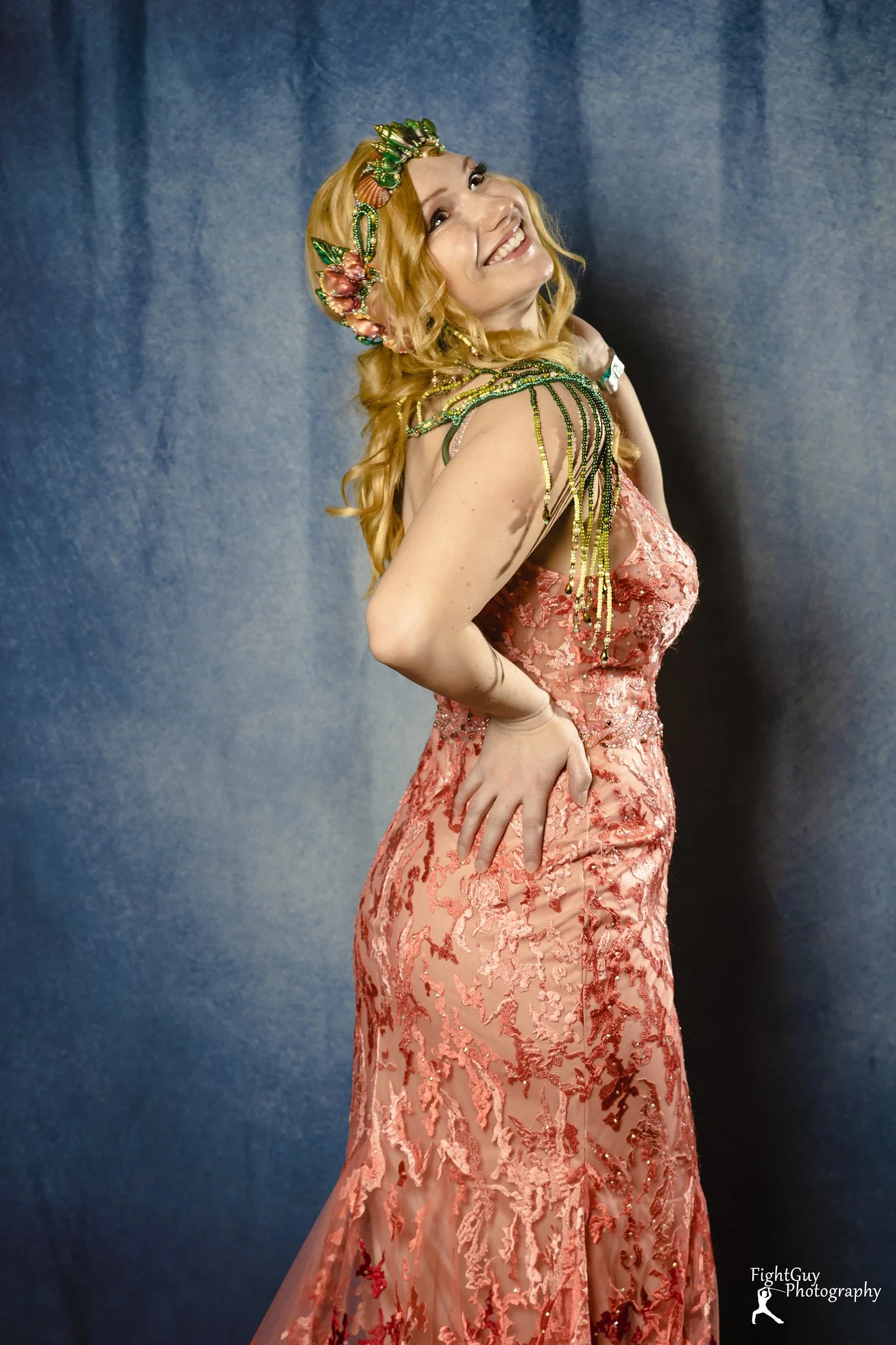 A woman with long, curly blonde hair wearing a pink lace gown with floral embroidery, standing against a blue backdrop, smiling and looking upward.