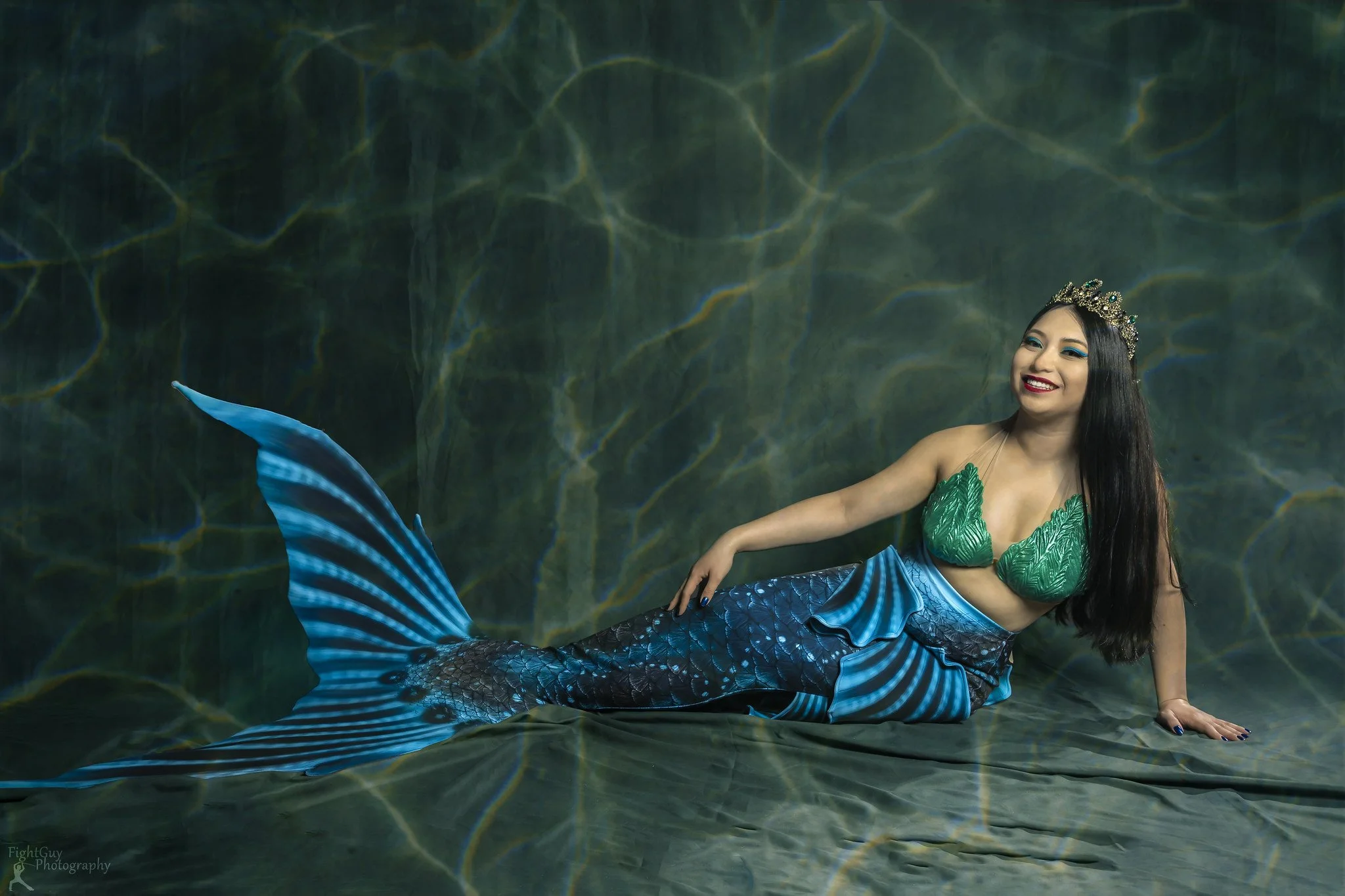 A woman dressed as a mermaid with a green top, blue tail, and a crown, smiling while lying on a green fabric background.